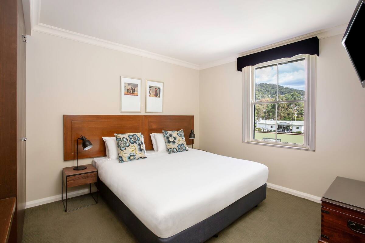 Un dormitorio ordenado con una cama grande, una mesita de noche, una lámpara y una ventana que ofrece vistas a un paisaje verde.