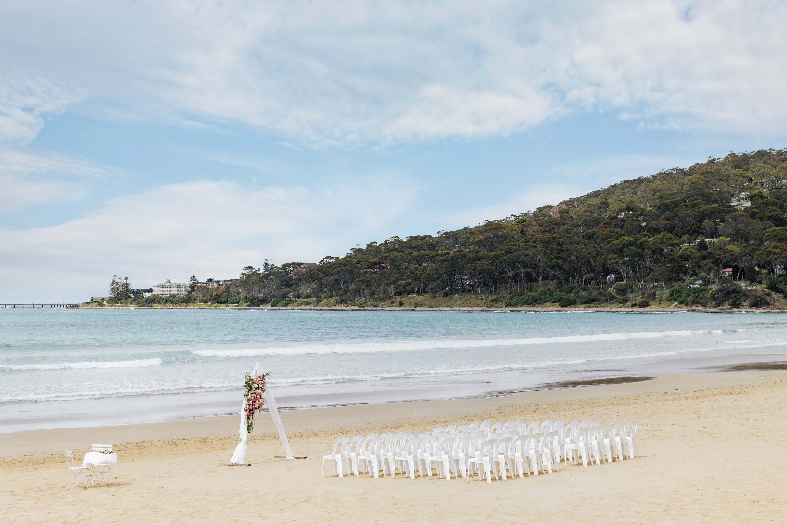 A beach wedding setup with white chairs and a floral arch.
