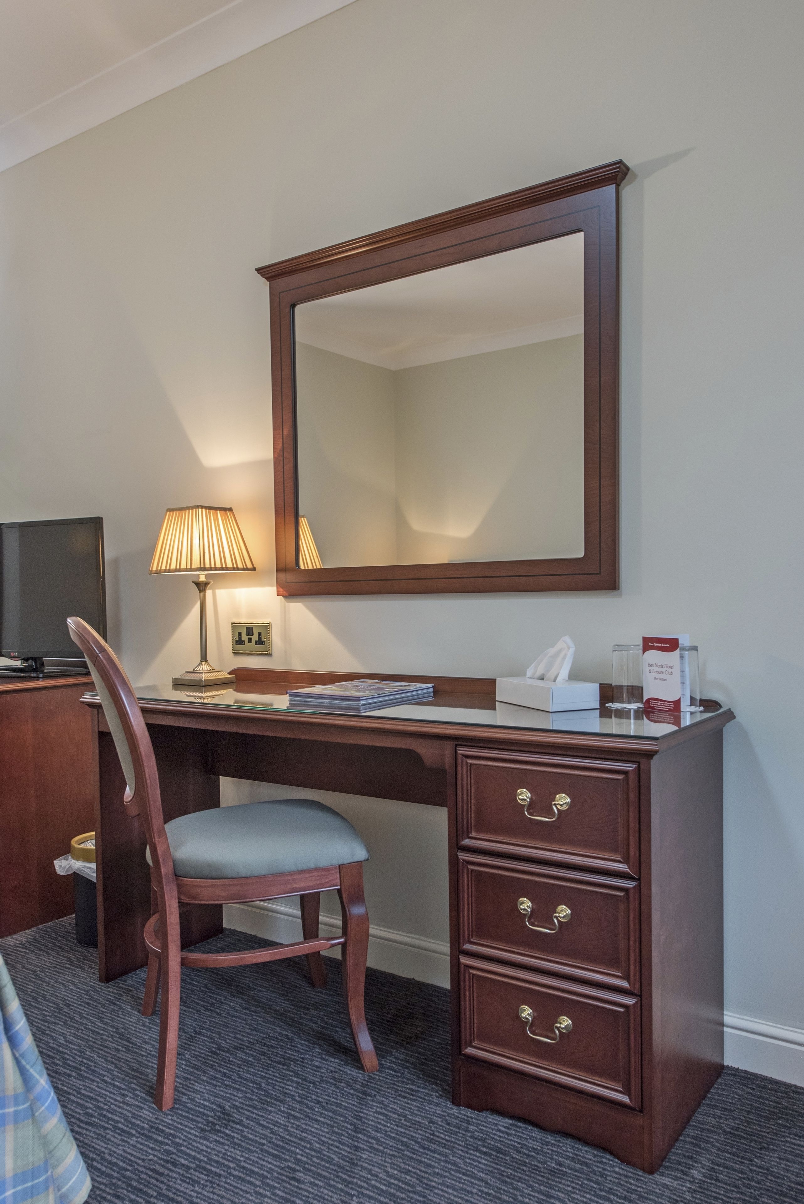 A hotel room with a wooden desk, a chair, a lamp, a mirror, and a telephone.
