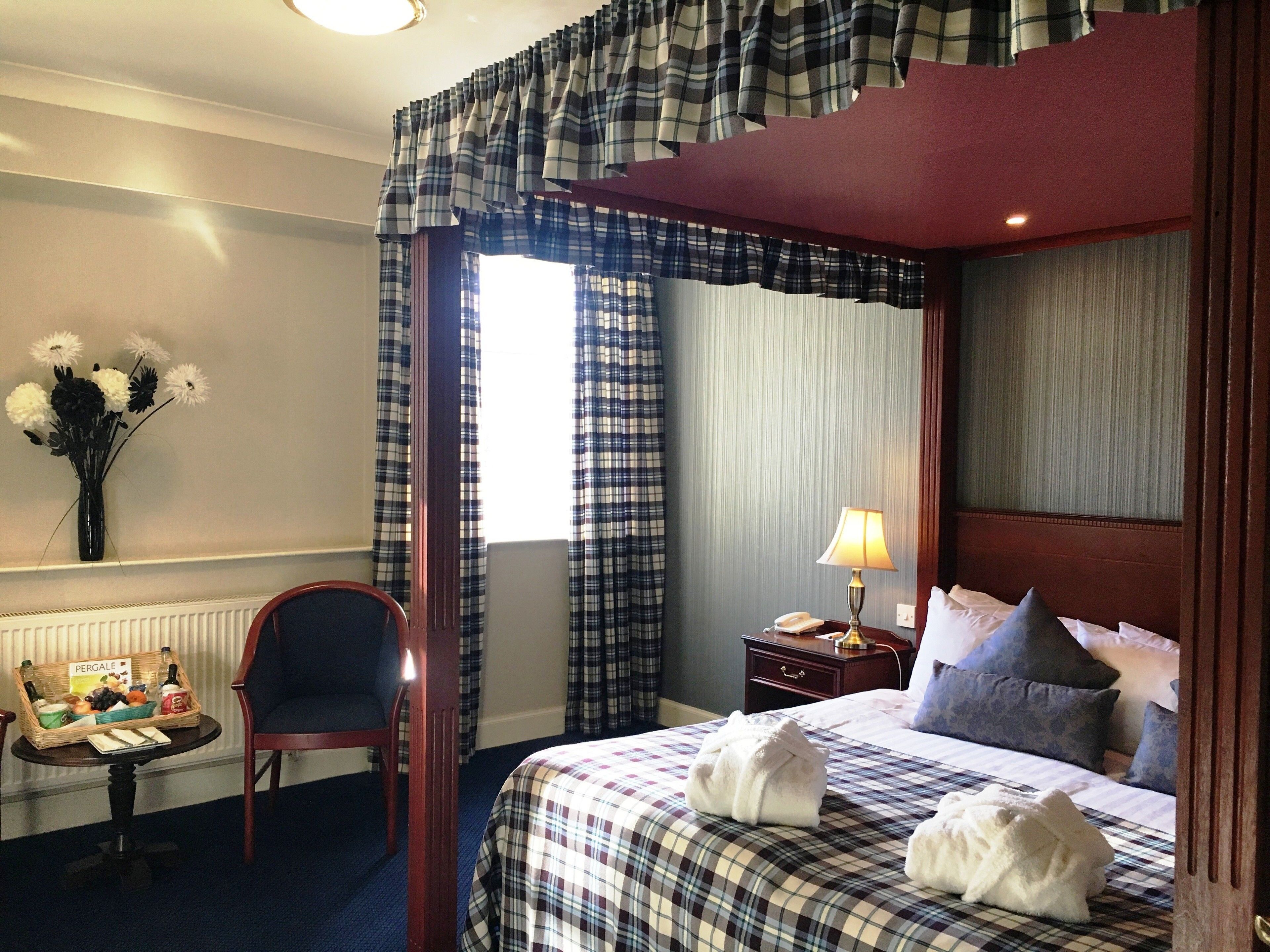 A hotel room with a bed, a chair, a nightstand, a lamp, a vase with flowers, and a window with curtains.