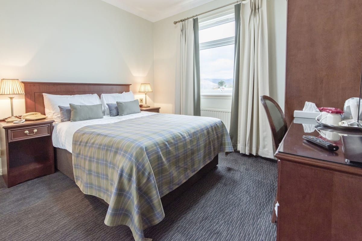 A hotel room with a bed, two bedside tables with lamps, a desk with a chair, and a window with curtains.
