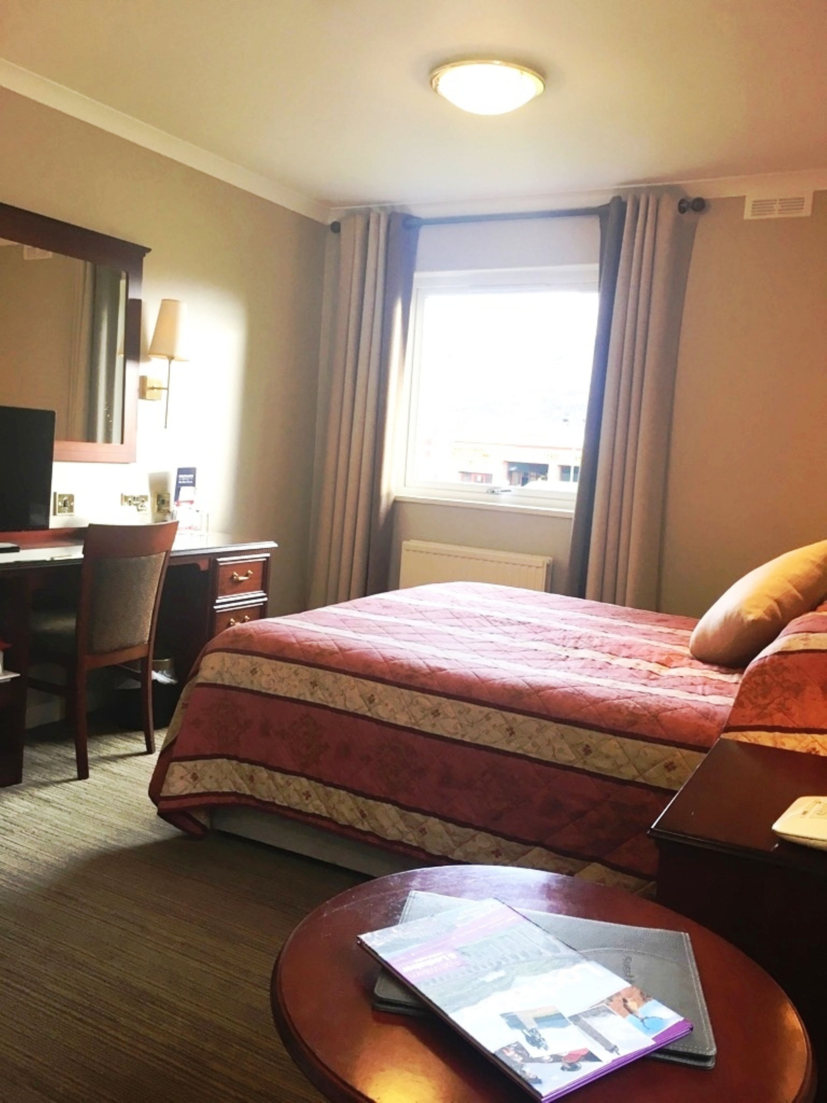 A hotel room with a bed, a desk, a chair, a lamp, and a window with curtains.