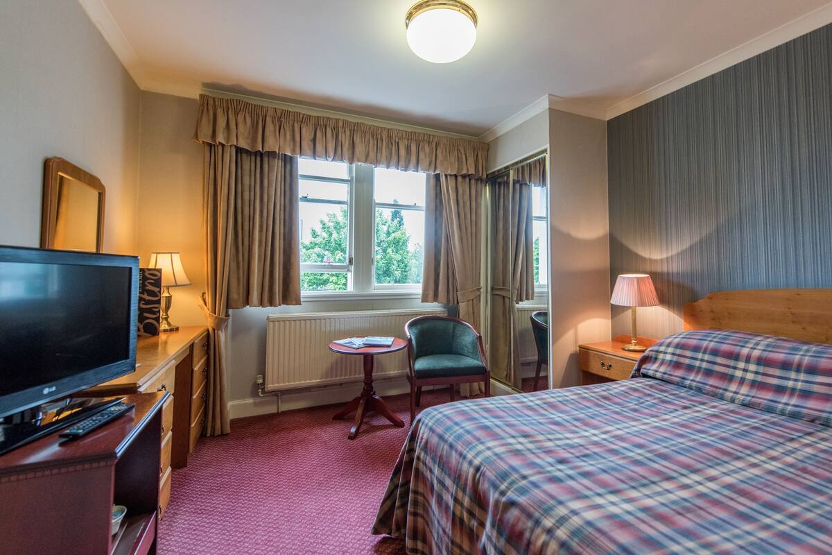 A hotel room with a bed, a TV, a desk with a lamp, a chair, and a window with curtains.