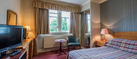 Habitación de hotel con cama, televisión, escritorio con lámpara, silla y ventana con cortinas.