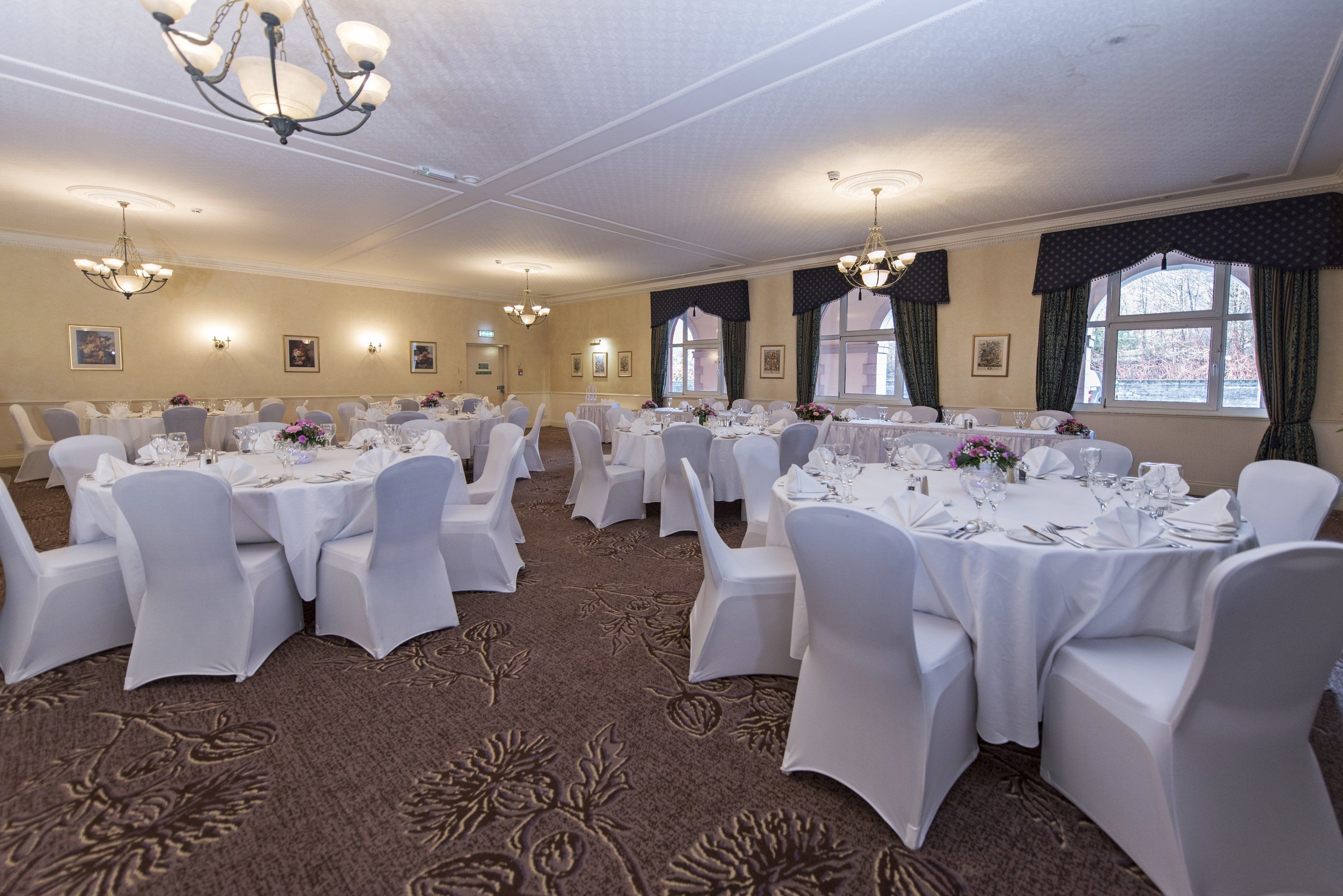A banquet hall with round tables, white tablecloths, and chairs, featuring chandeliers and framed pictures on the walls.