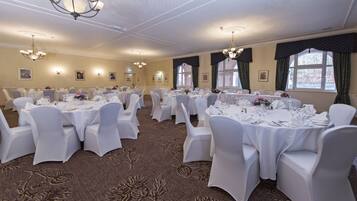 A banquet hall with round tables, white tablecloths, and chairs, featuring chandeliers and framed pictures on the walls.