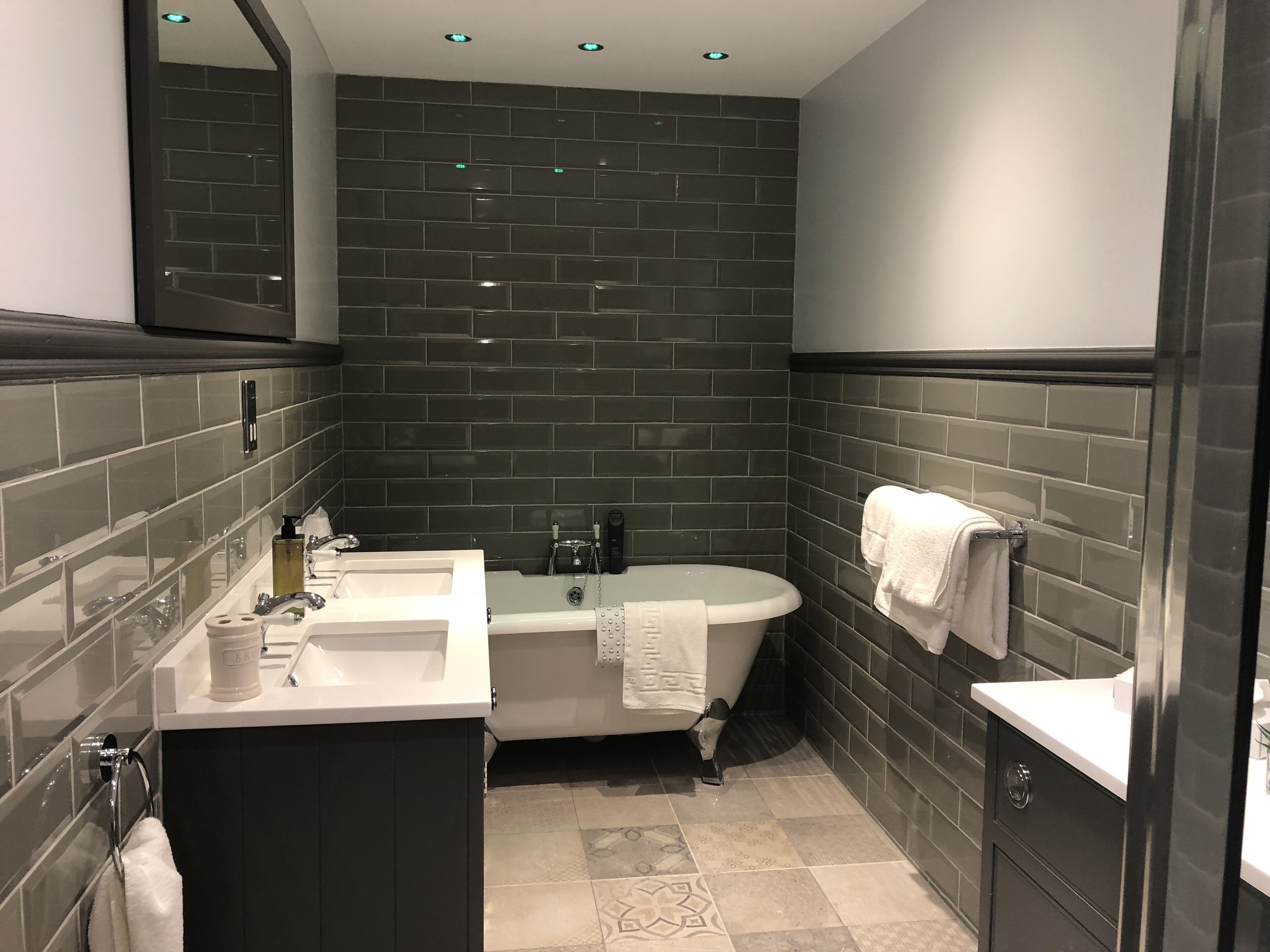 Un baño moderno con paredes de azulejos oscuros, un lavabo blanco, una bañera y un espejo sobre el lavabo.