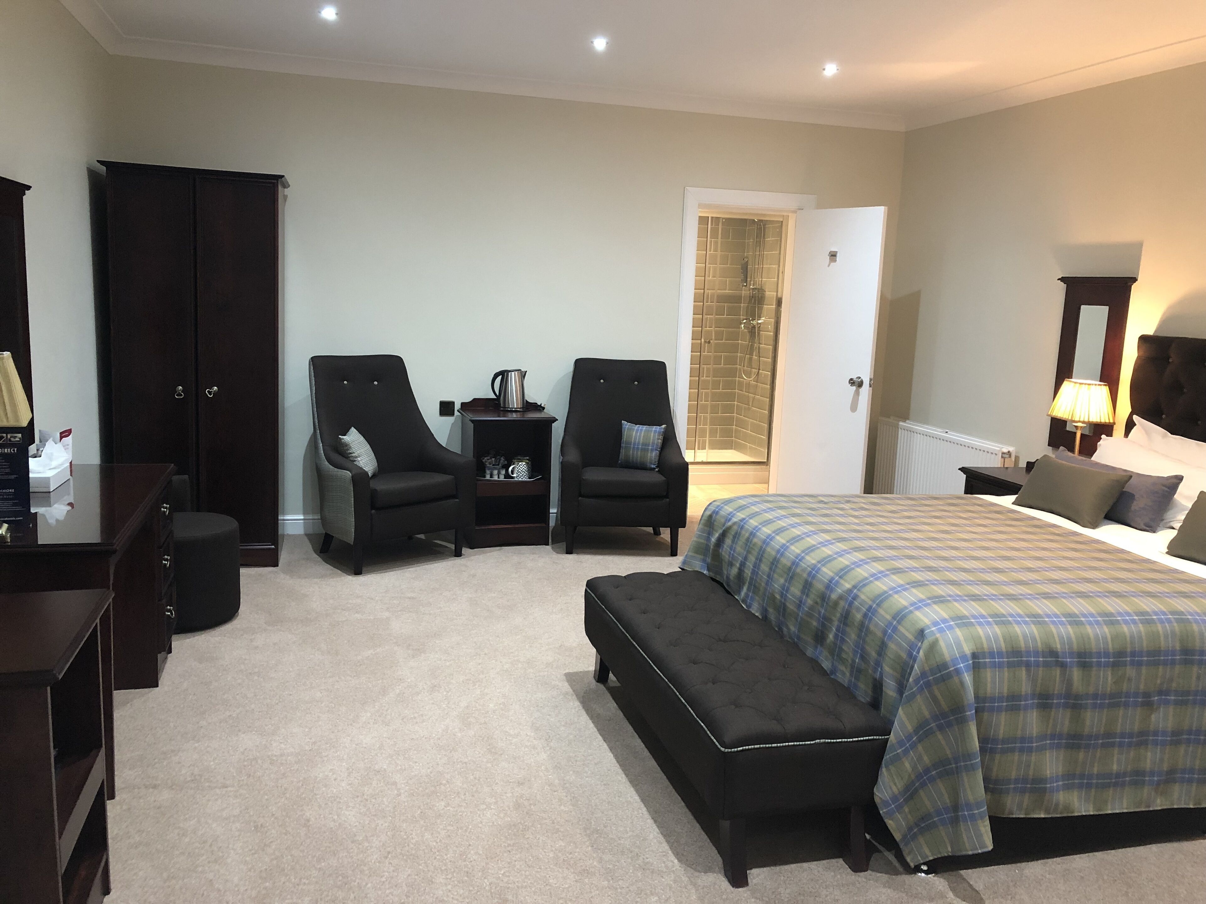 A bedroom with a large bed, two armchairs, a bench, a dresser, a wardrobe, and a shower area.