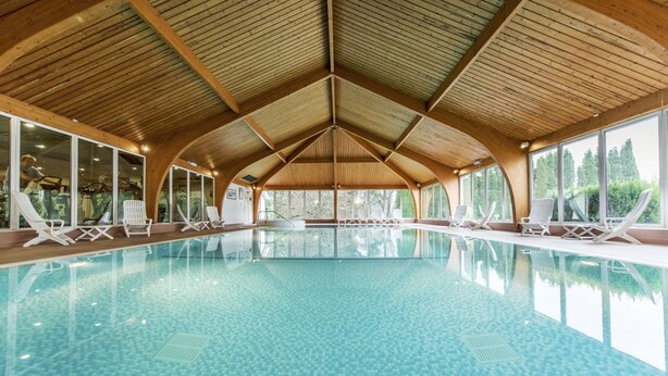 Une piscine intérieure avec des poutres en bois, des parois en verre transparent et des chaises longues blanches.