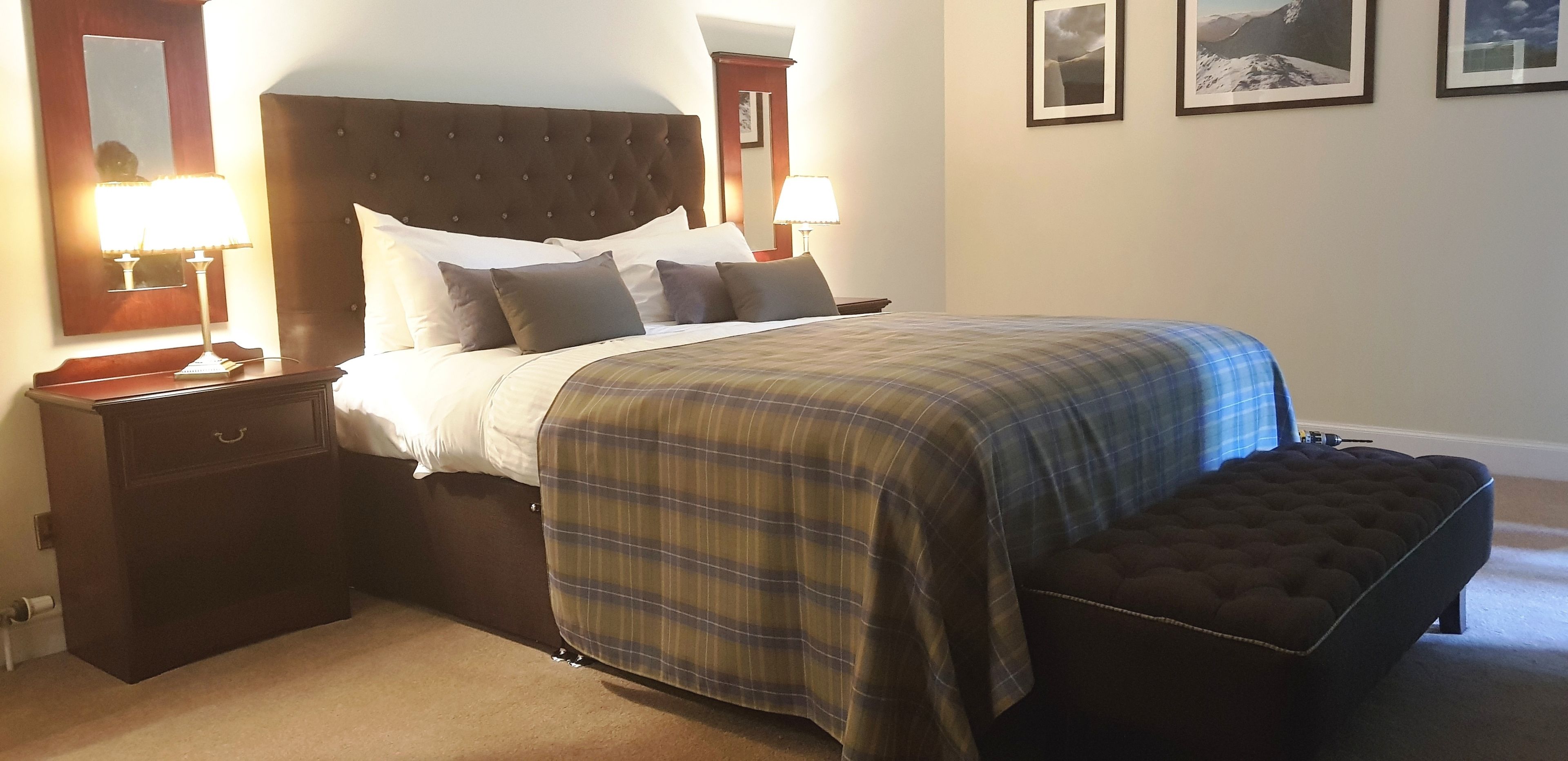 A neatly made bed with a plaid bedspread, two bedside lamps, a nightstand, and a bench.