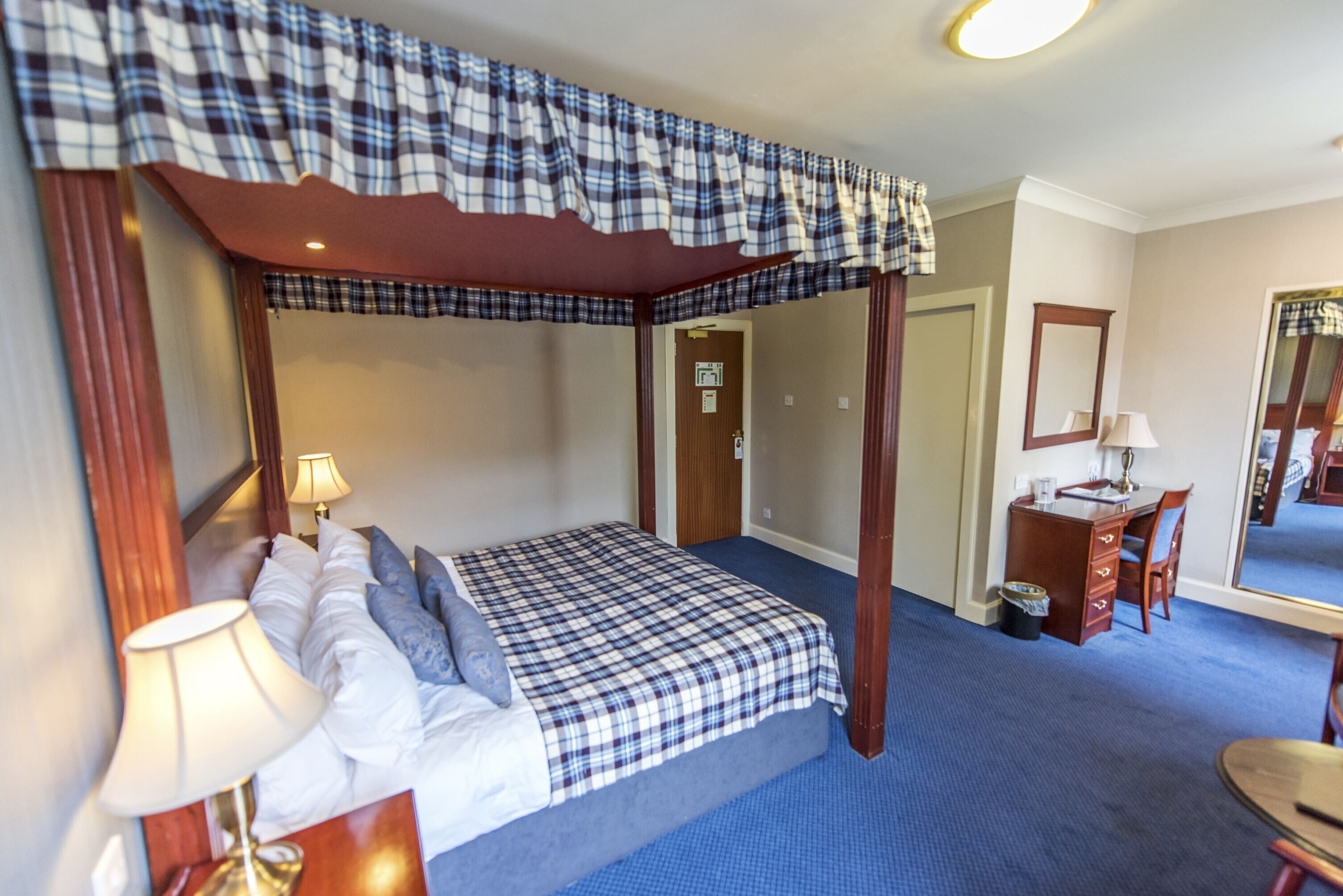 A four-poster bed with a plaid bedspread, a desk with a chair, a mirror, and a lamp.
