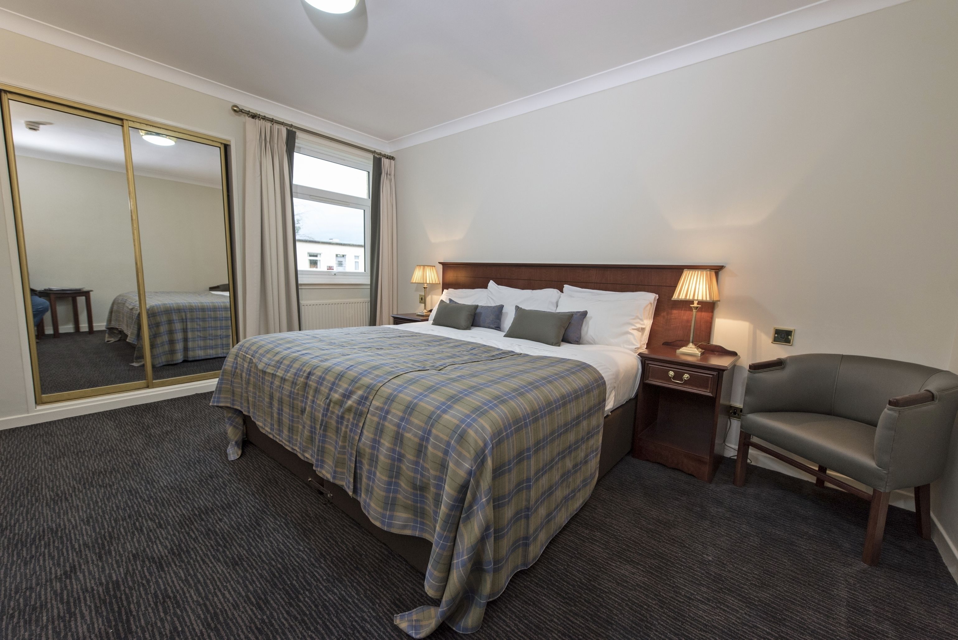 A hotel room with a large bed, a chair, a nightstand, a mirror, and a window with curtains.