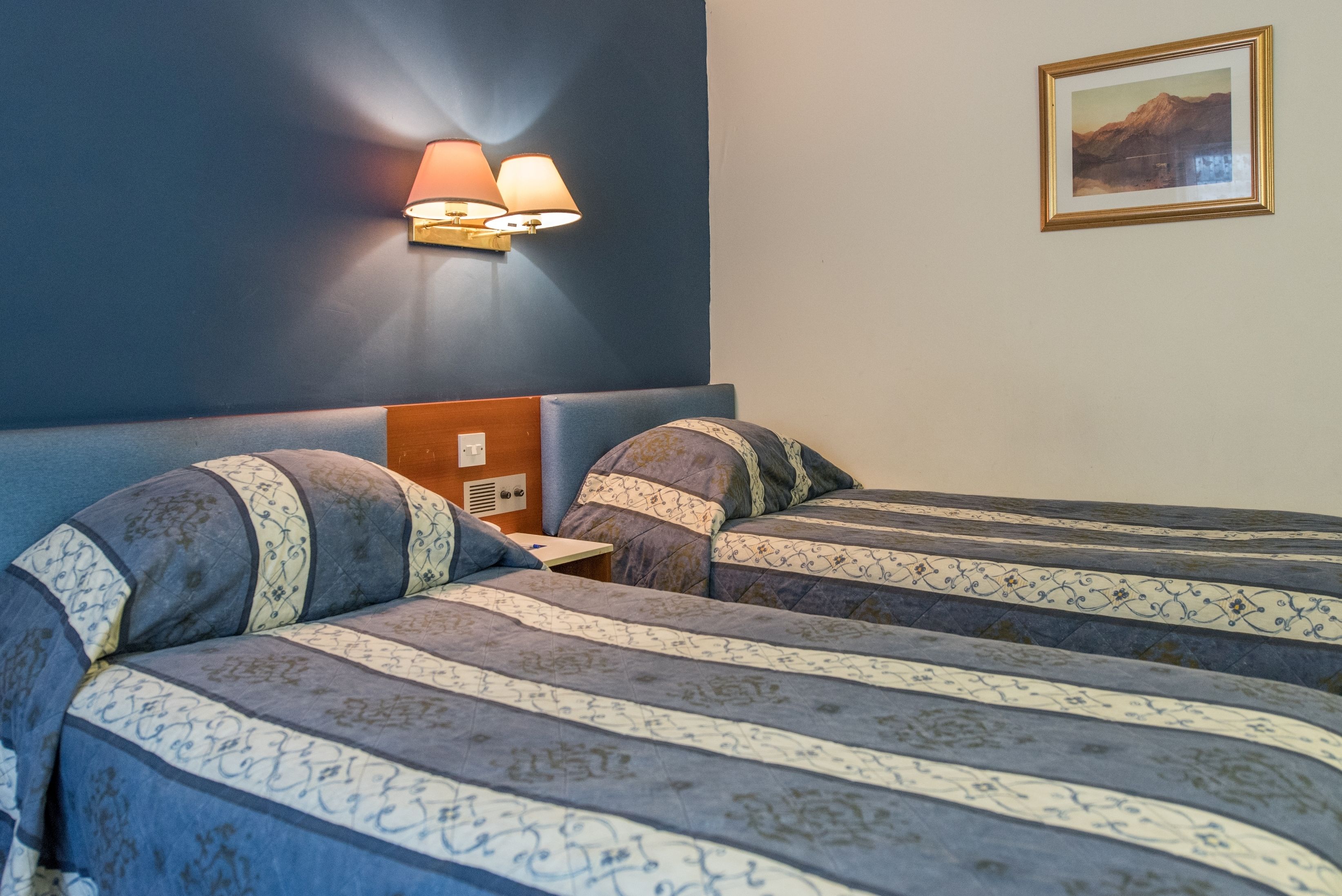 A hotel room with two beds, a nightstand, a wall-mounted lamp, and a framed picture on the wall.