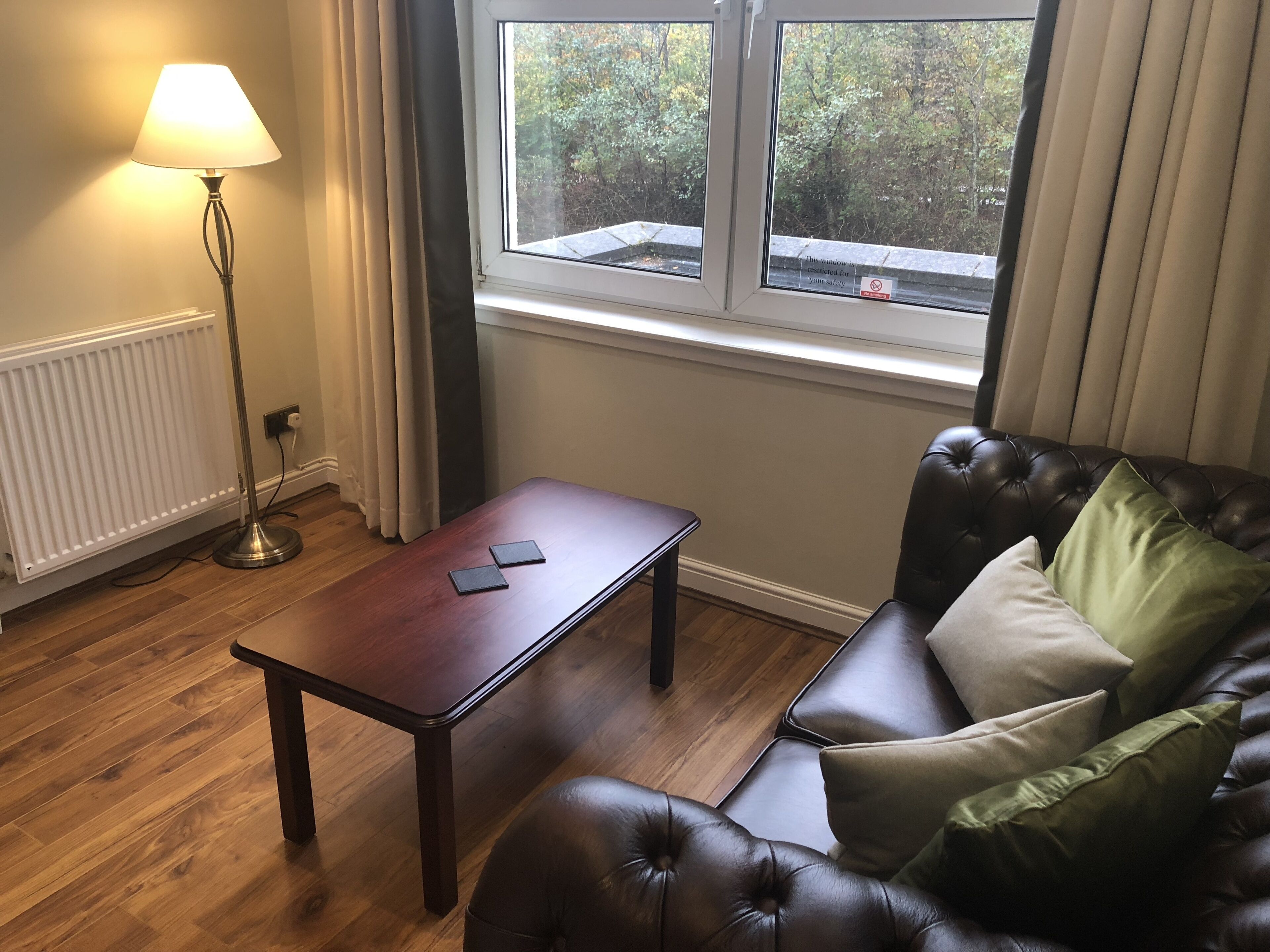 Ein Zimmer mit Holzboden, einem Ledersofa mit Kissen, einem kleinen Holztisch, einer Stehlampe und einem Fenster mit Blick auf Bäume.