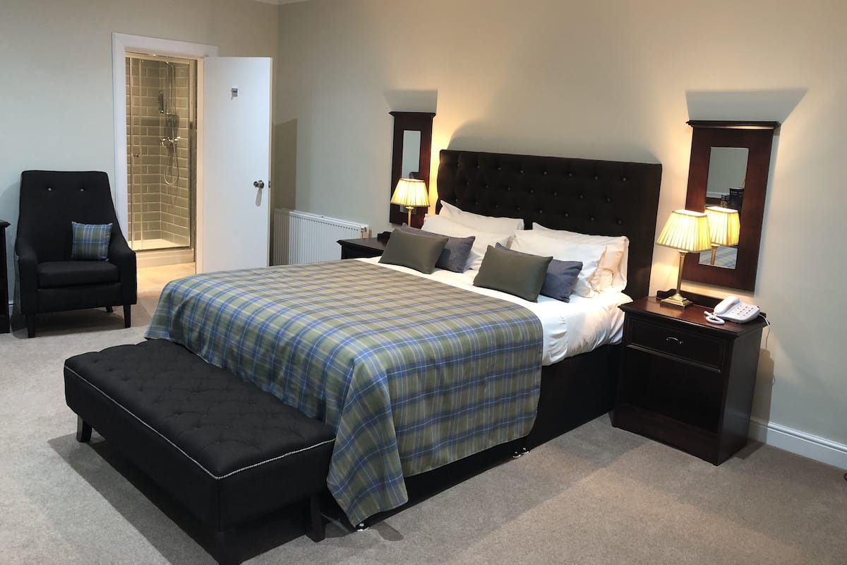 A bedroom with a large bed, a bench, a chair, a bedside table with a lamp, and a door leading to a bathroom.