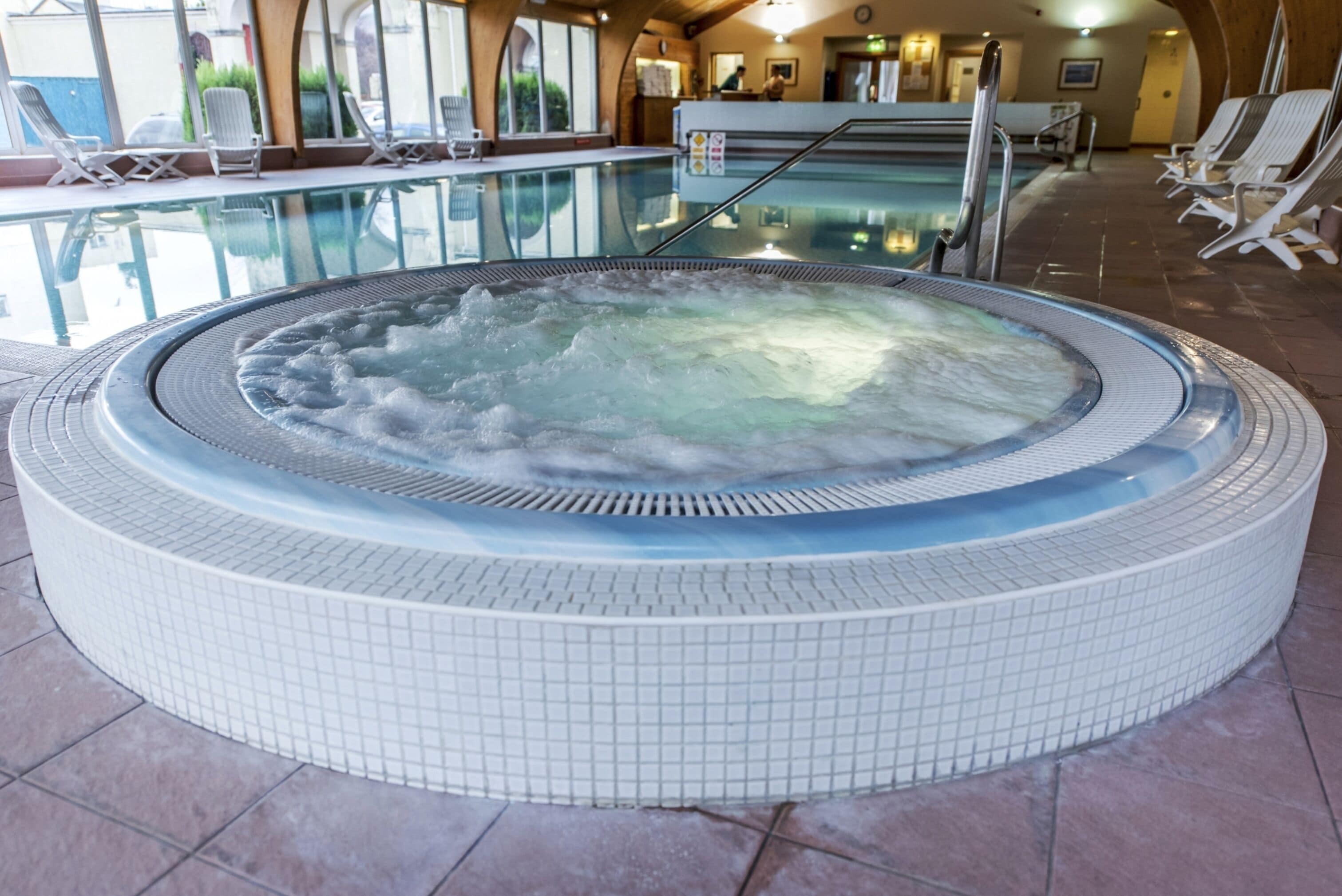 A hot tub with a tiled edge, surrounded by lounge chairs and a pool.