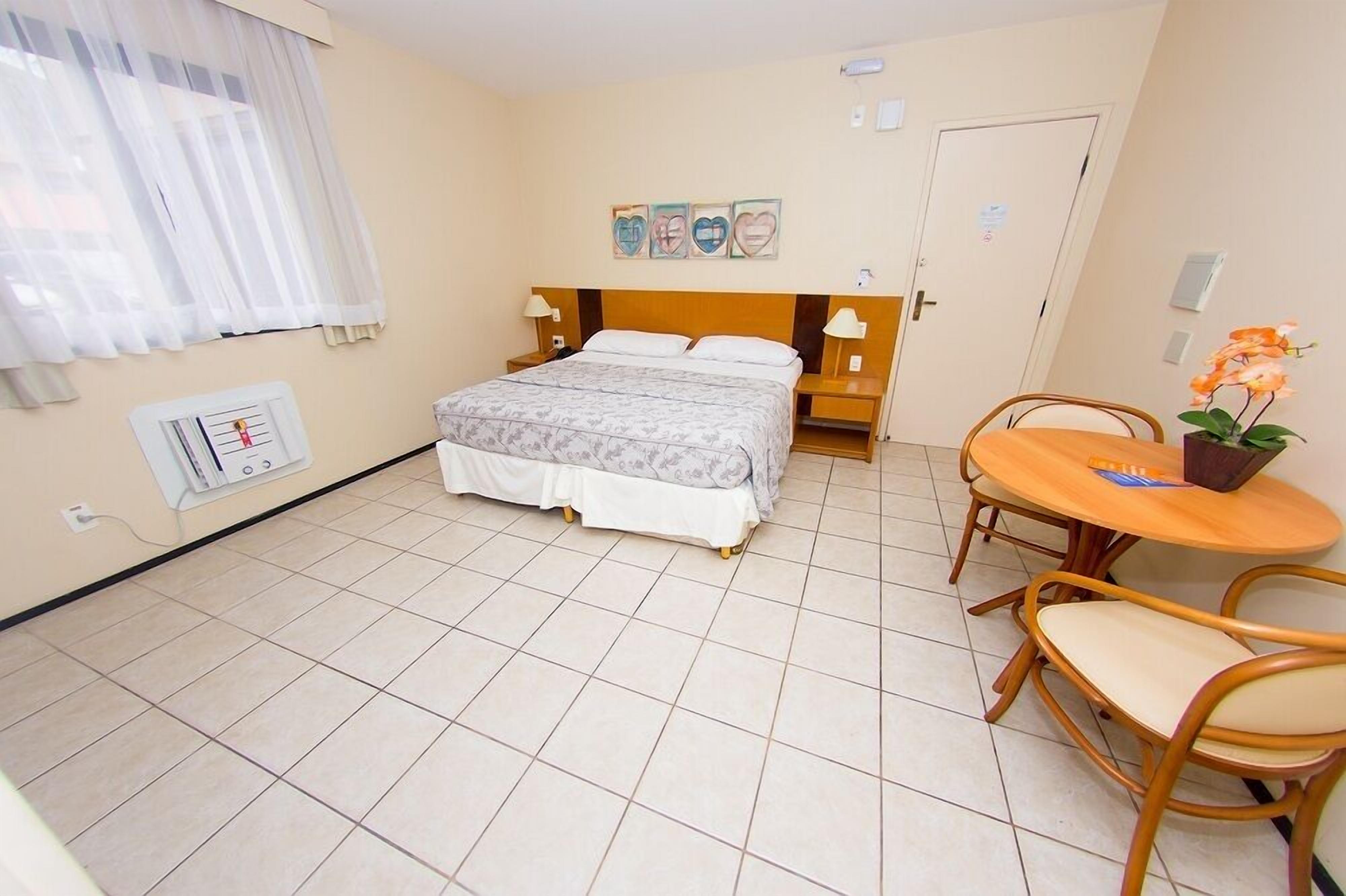 A hotel room with a bed, bedside tables, a small round table with chairs, a window with sheer curtains, and a wall-mounted air conditioning unit.