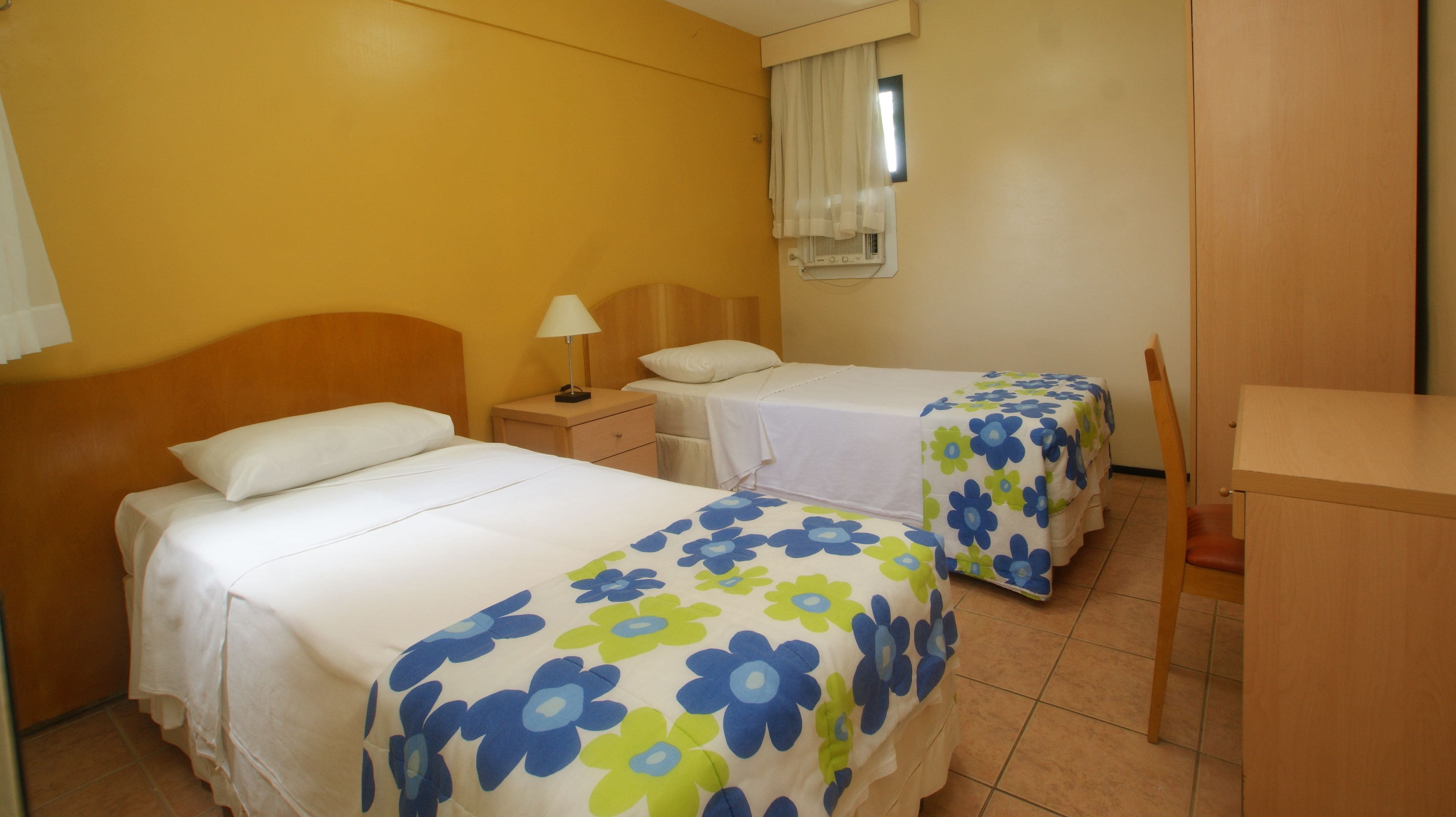 Ein Hotelzimmer mit zwei Betten, einem Schreibtisch, einem Stuhl, einem Nachttisch, einer Lampe und einem Fenster mit Vorhängen.