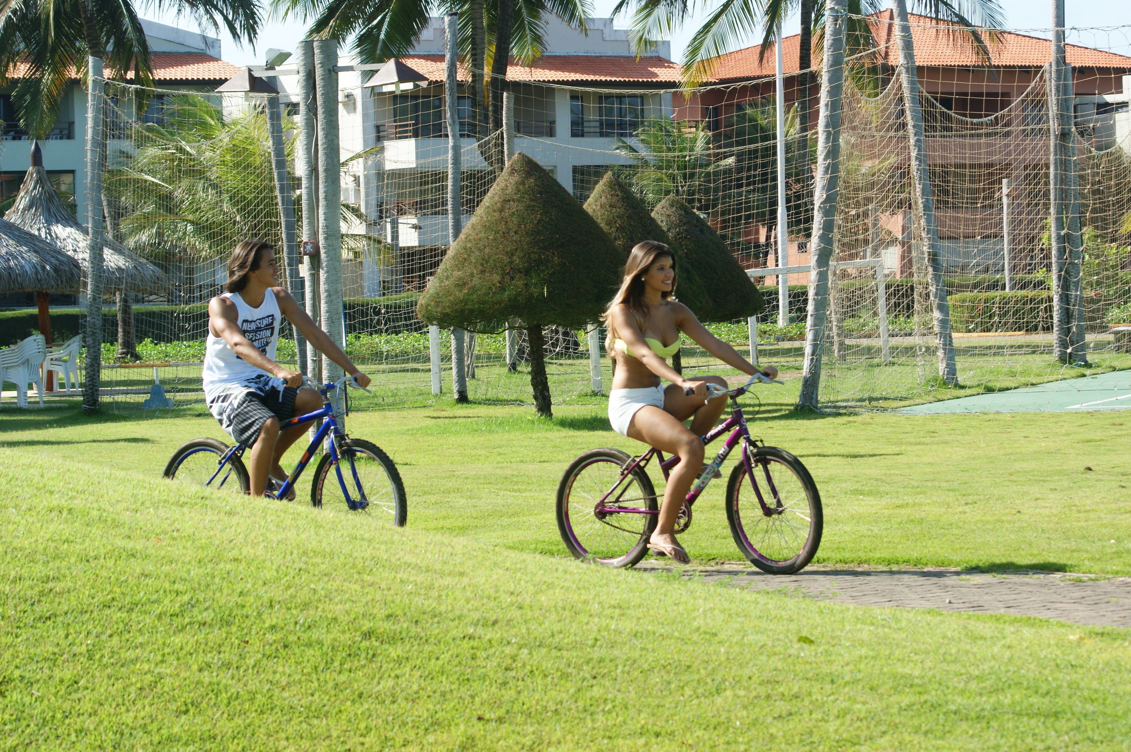 Dos personas andan en bicicleta por un césped con una cerca y árboles al fondo.