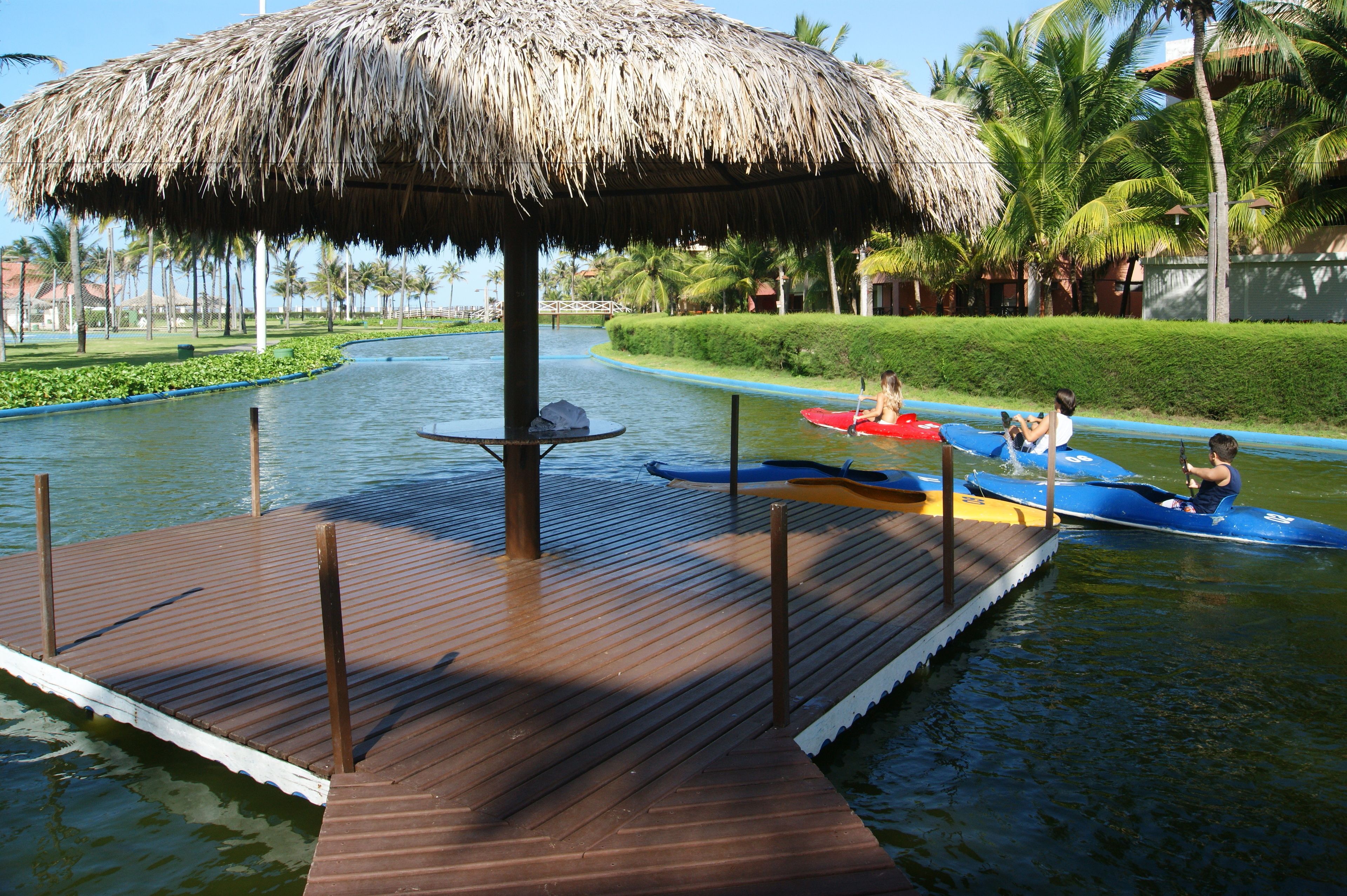 A wooden dock with a thatched roof, a water channel, and kayaks.