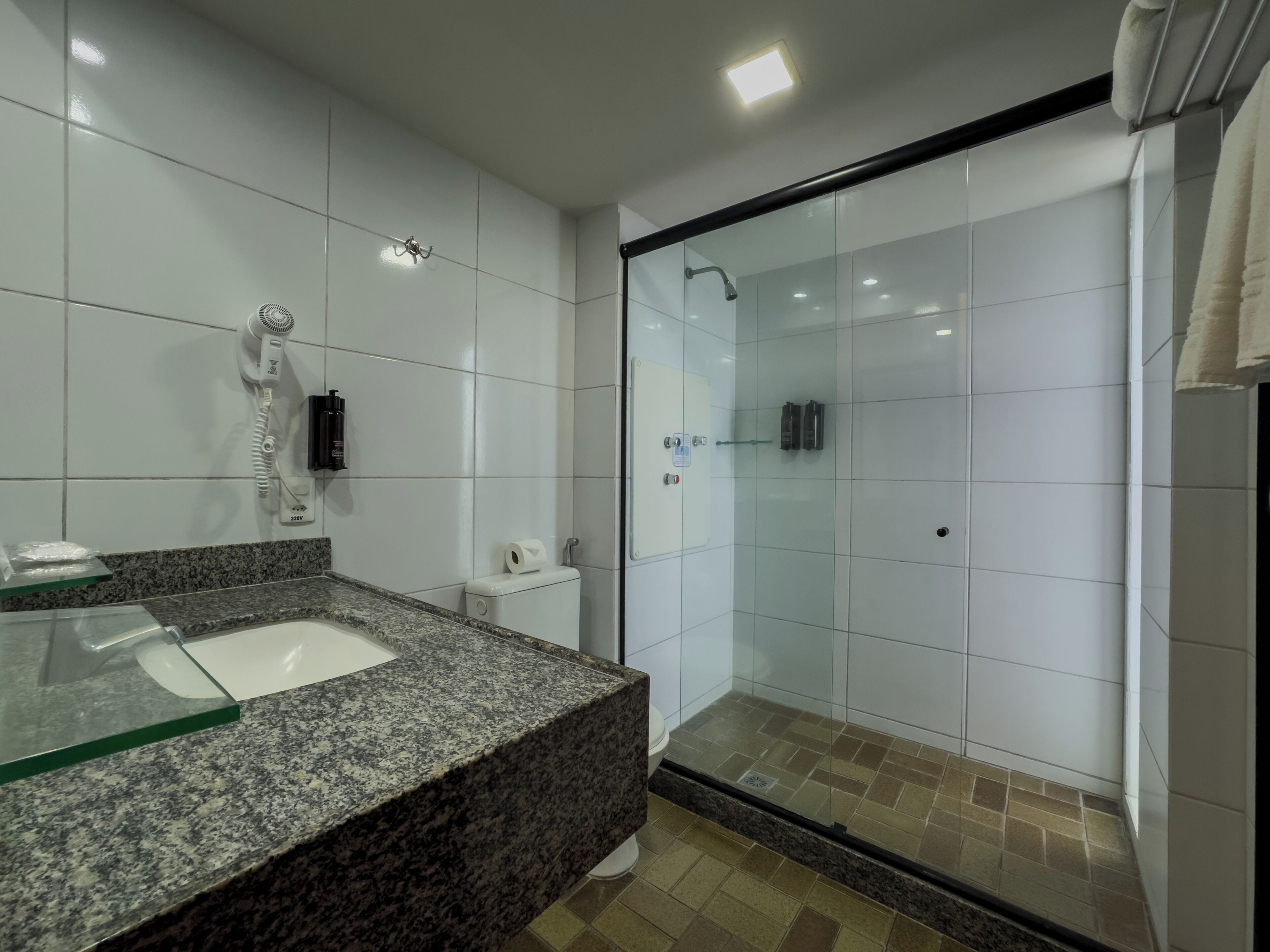 Un baño moderno con lavamanos de granito, azulejos blancos, cabina de ducha con mampara de vidrio y un secador de pelo montado en la pared.