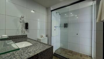 A modern bathroom with a granite countertop sink, white tiles, a glass shower enclosure, and a hairdryer mounted on the wall.