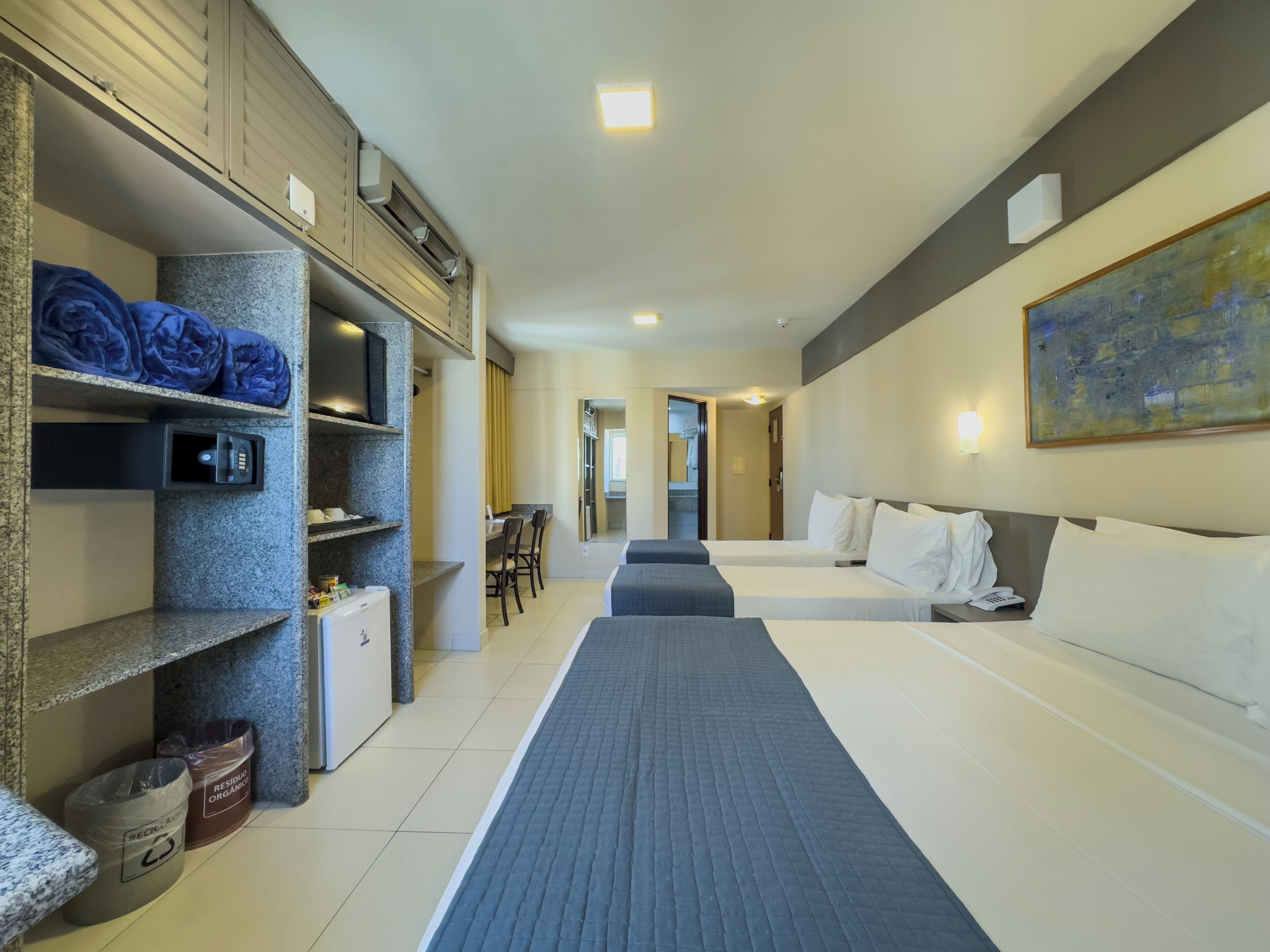 A hotel room with two beds, a desk, a trash bin, and a painting on the wall.