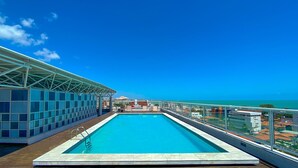 Una piscina en la azotea con agua azul cristalina, rodeada por una barandilla de cristal, y con vistas al océano y al paisaje urbano.