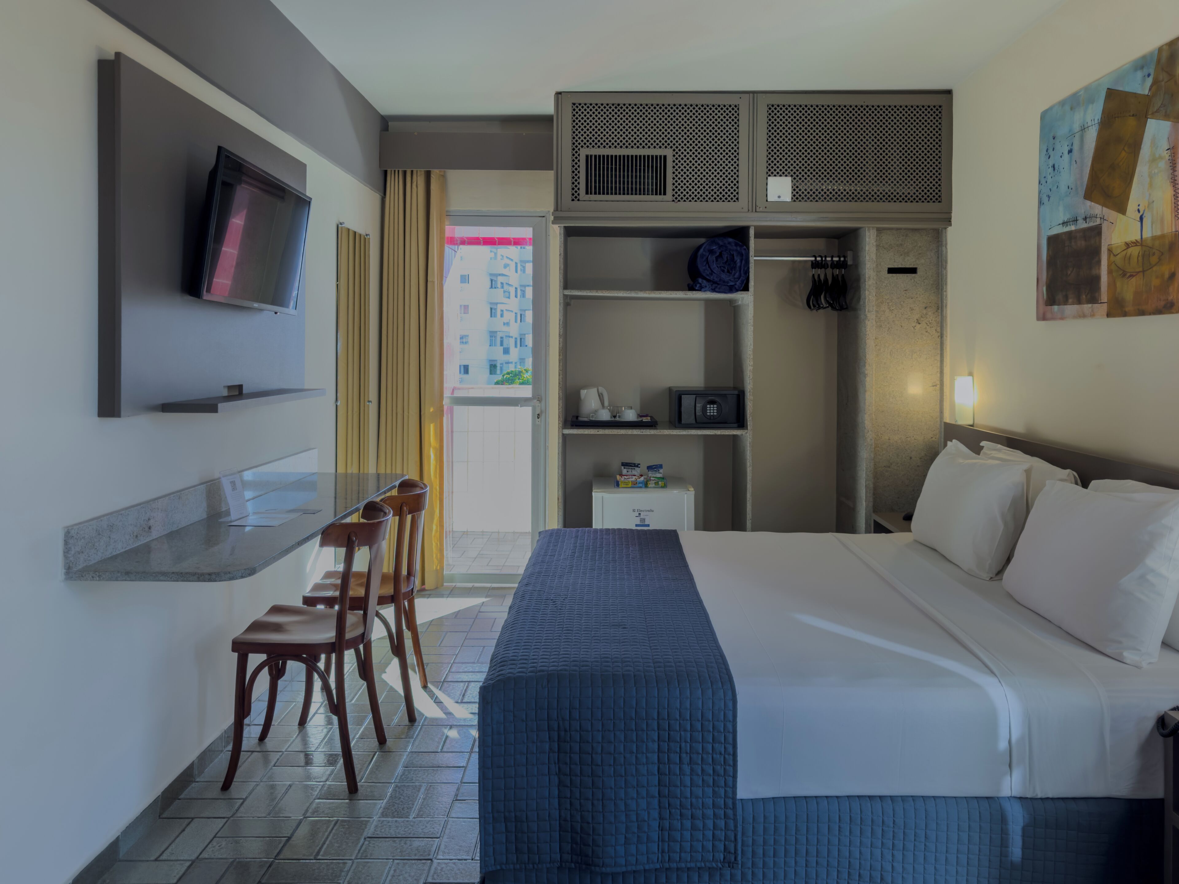 A hotel room with a bed, a desk with chairs, a television, a wardrobe, and a painting on the wall.