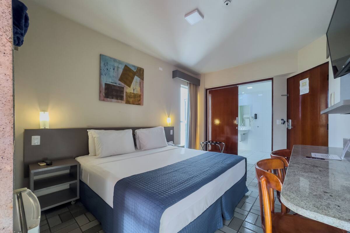 Een hotelkamer met een bed, nachtkastje, lamp, kunstwerk en een klein kitchenette.