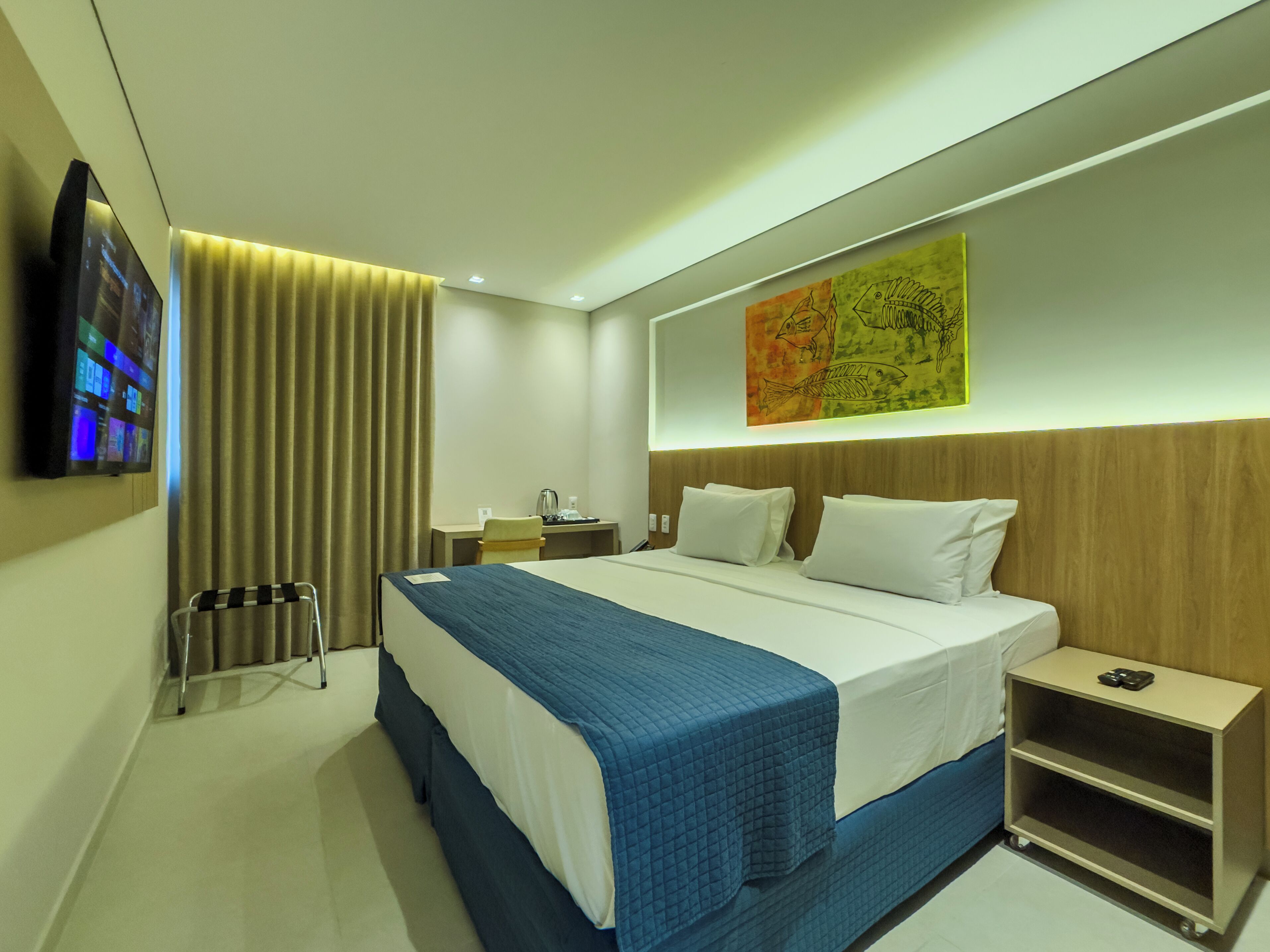 A modern hotel room with a large bed, a desk, a chair, a TV, and a wall art piece.