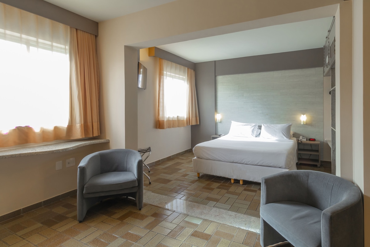A hotel room with a bed, two chairs, a desk, and a window with curtains.