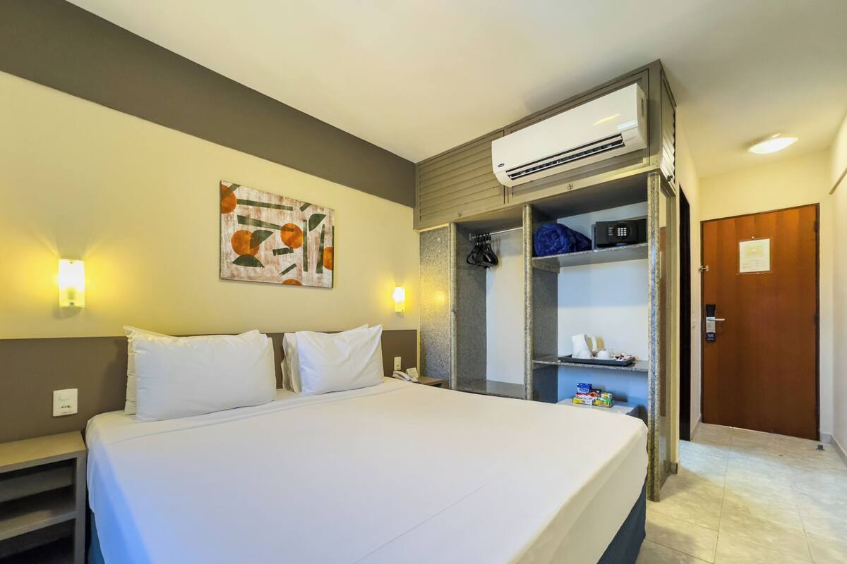Een hotelkamer met een bed, een airconditioning, een kunstwerk aan de muur, een kapstok en een deur.