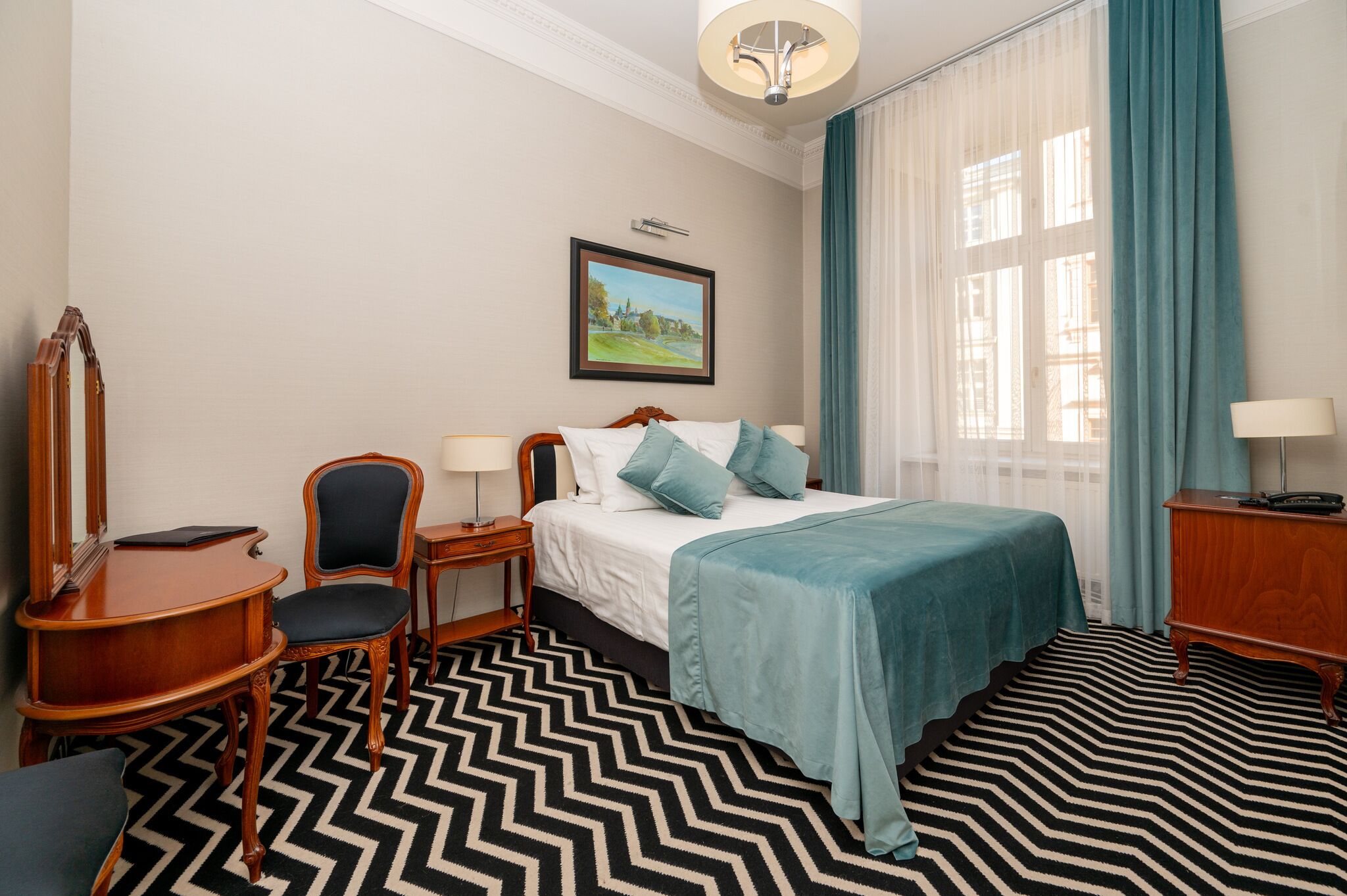 A hotel room with a bed, a desk, a chair, a mirror, a picture on the wall, and a window with curtains.