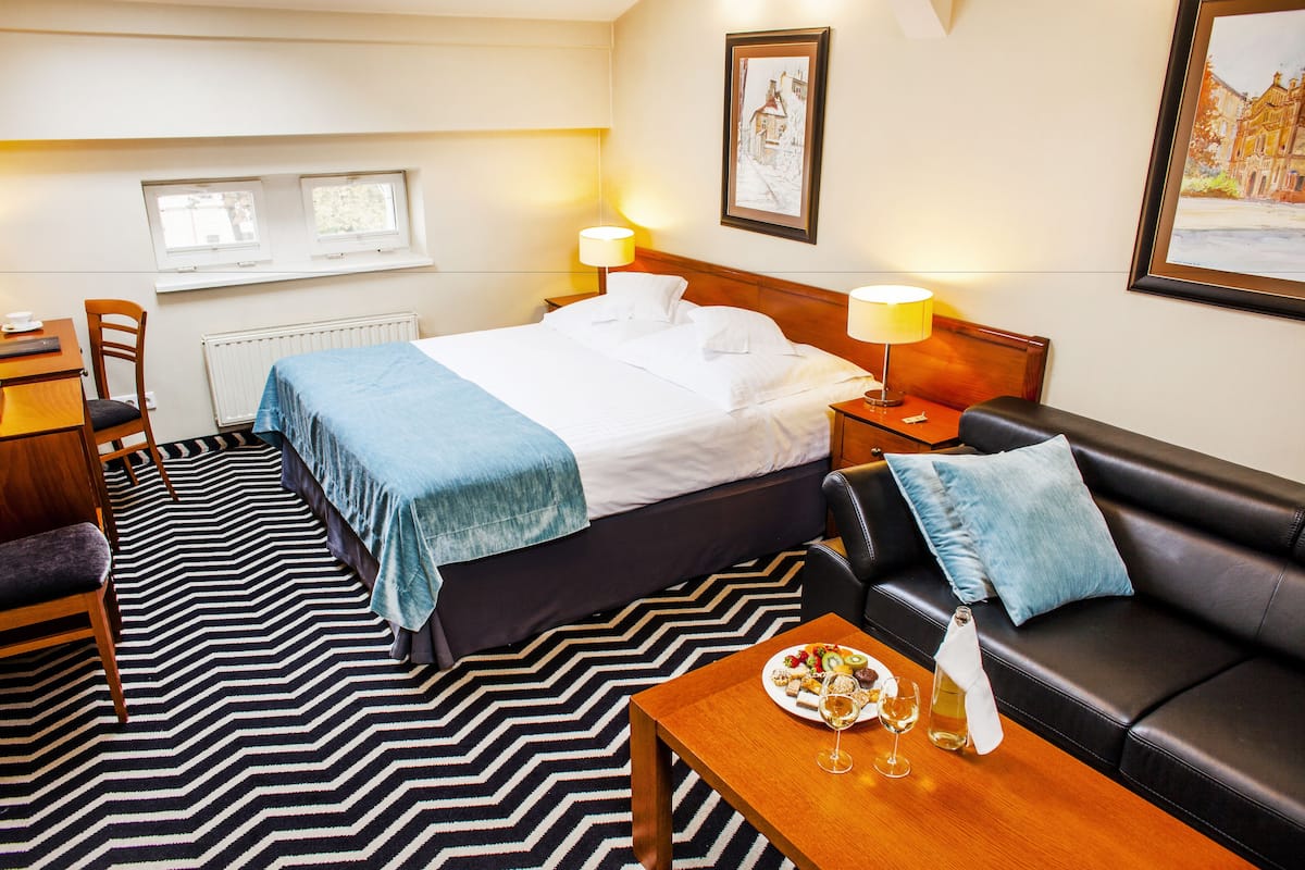 Ein Hotelzimmer mit einem Bett, einem Sofa, einem Esstisch mit Speisen und Getränken, einem Sessel, einem Schreibtisch und zwei Fenstern.