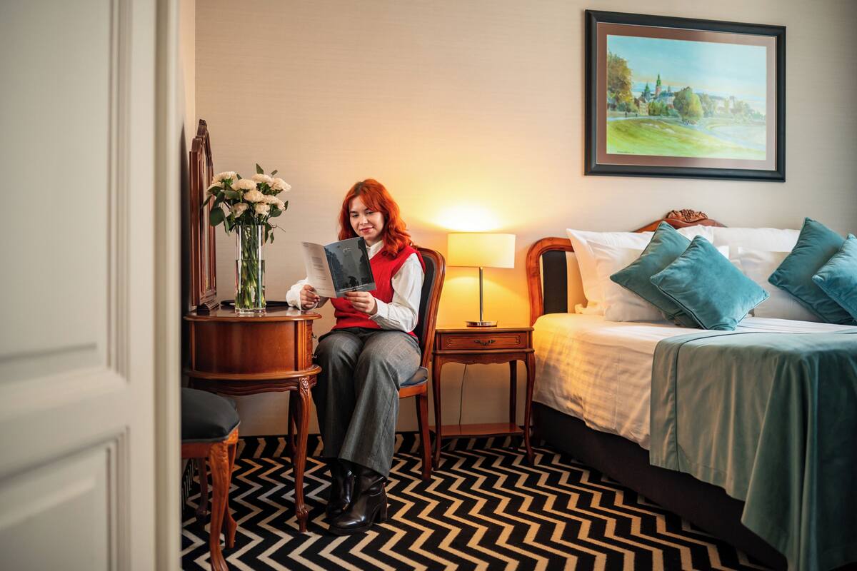 Ein Hotelzimmer mit einem Bett, einem Nachttisch mit Lampe, einem Sessel und einem Schreibtisch mit Blumenvase.