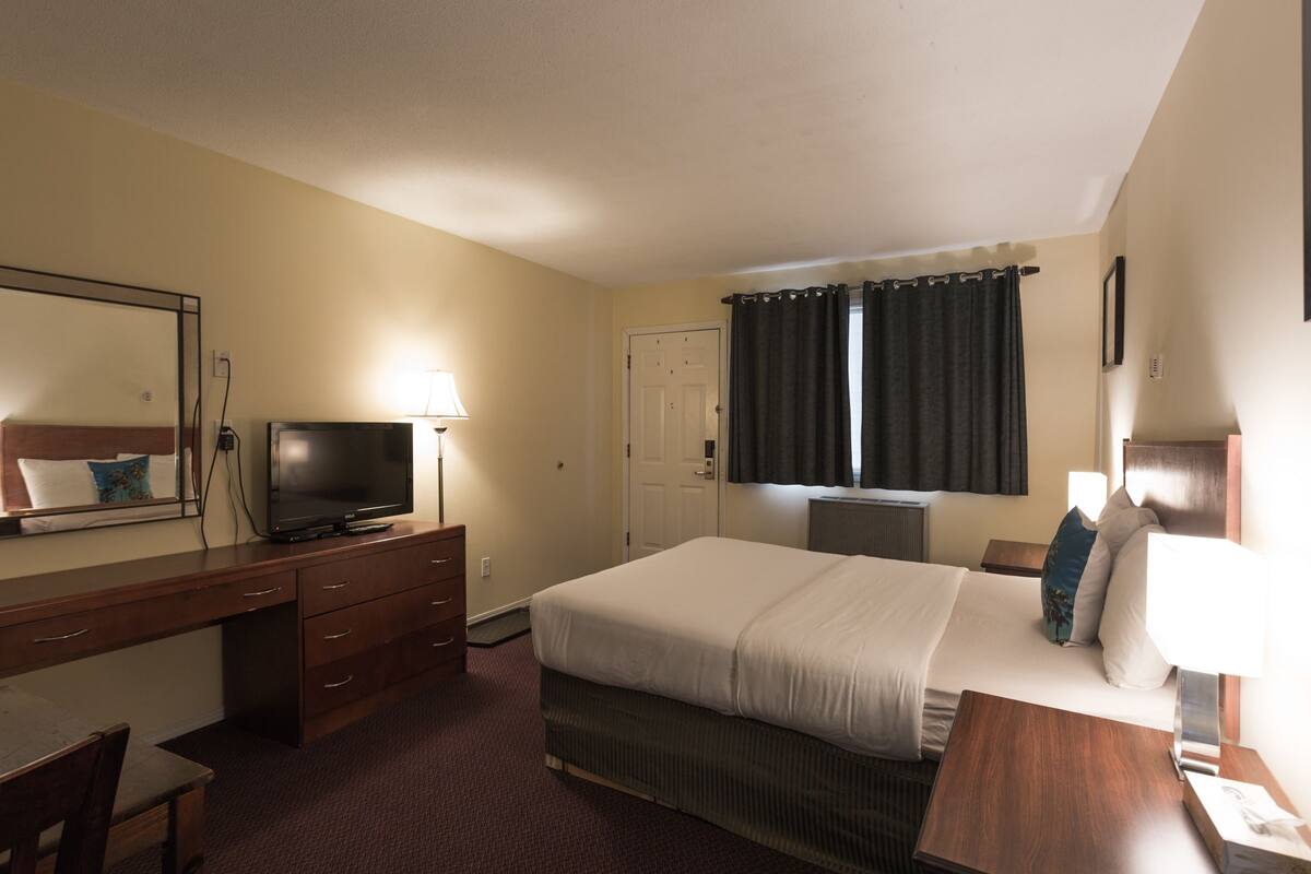 Una habitación de hotel con cama, escritorio, televisor, cómoda y ventana con cortinas.
