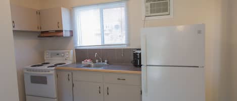 A compact kitchen with white appliances, a window with blinds, and a vent on the wall.