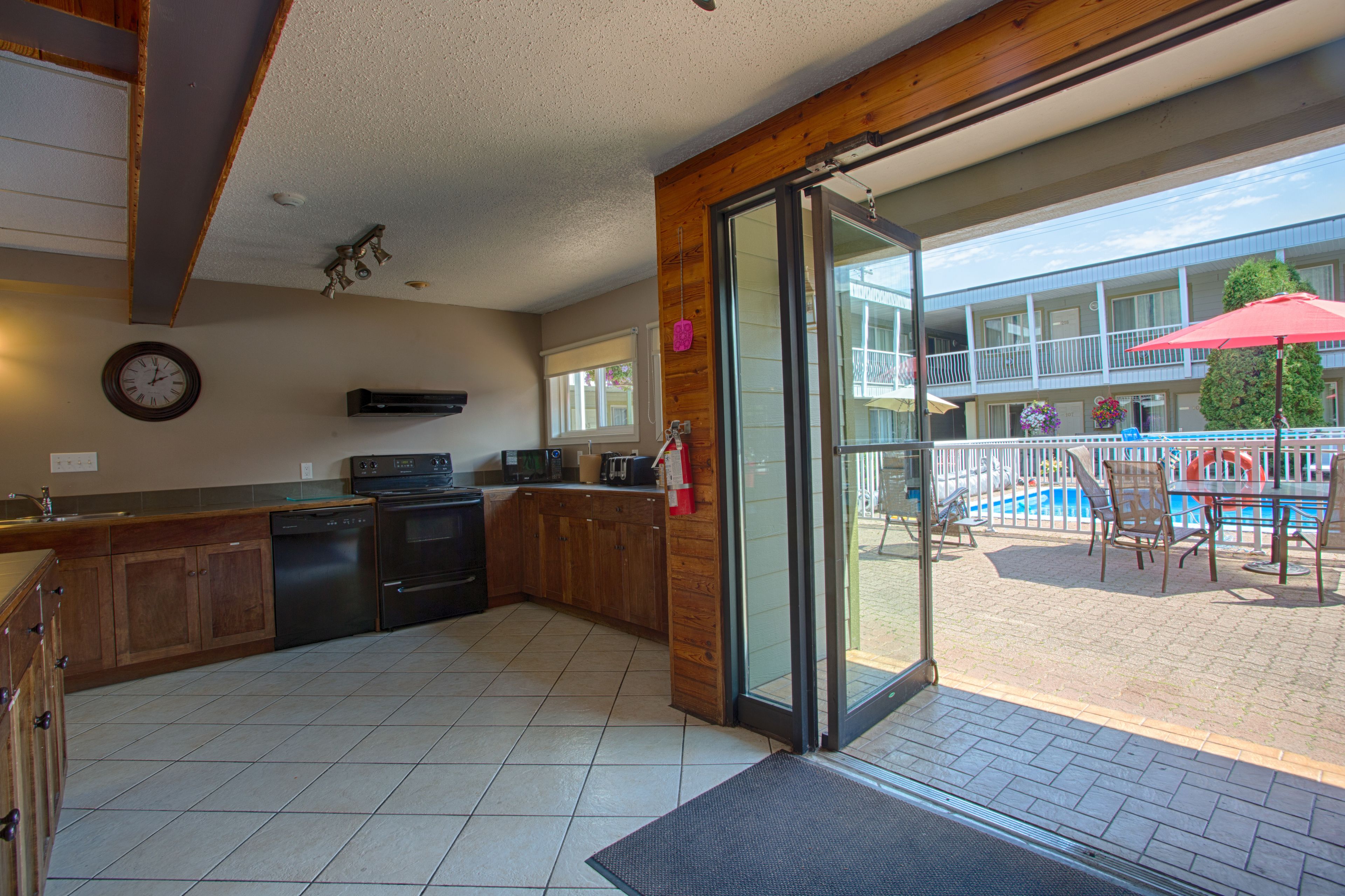 Una cocina con armarios de madera, lavavajillas y un amplio ventanal corredera que da a un patio exterior con piscina y sillas.
