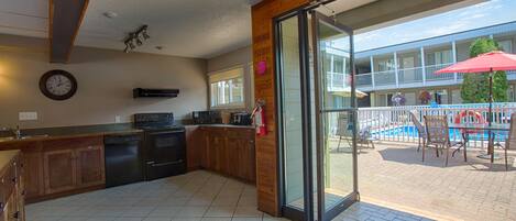 Una cocina con armarios de madera, lavavajillas y un amplio ventanal corredera que da a un patio exterior con piscina y sillas.