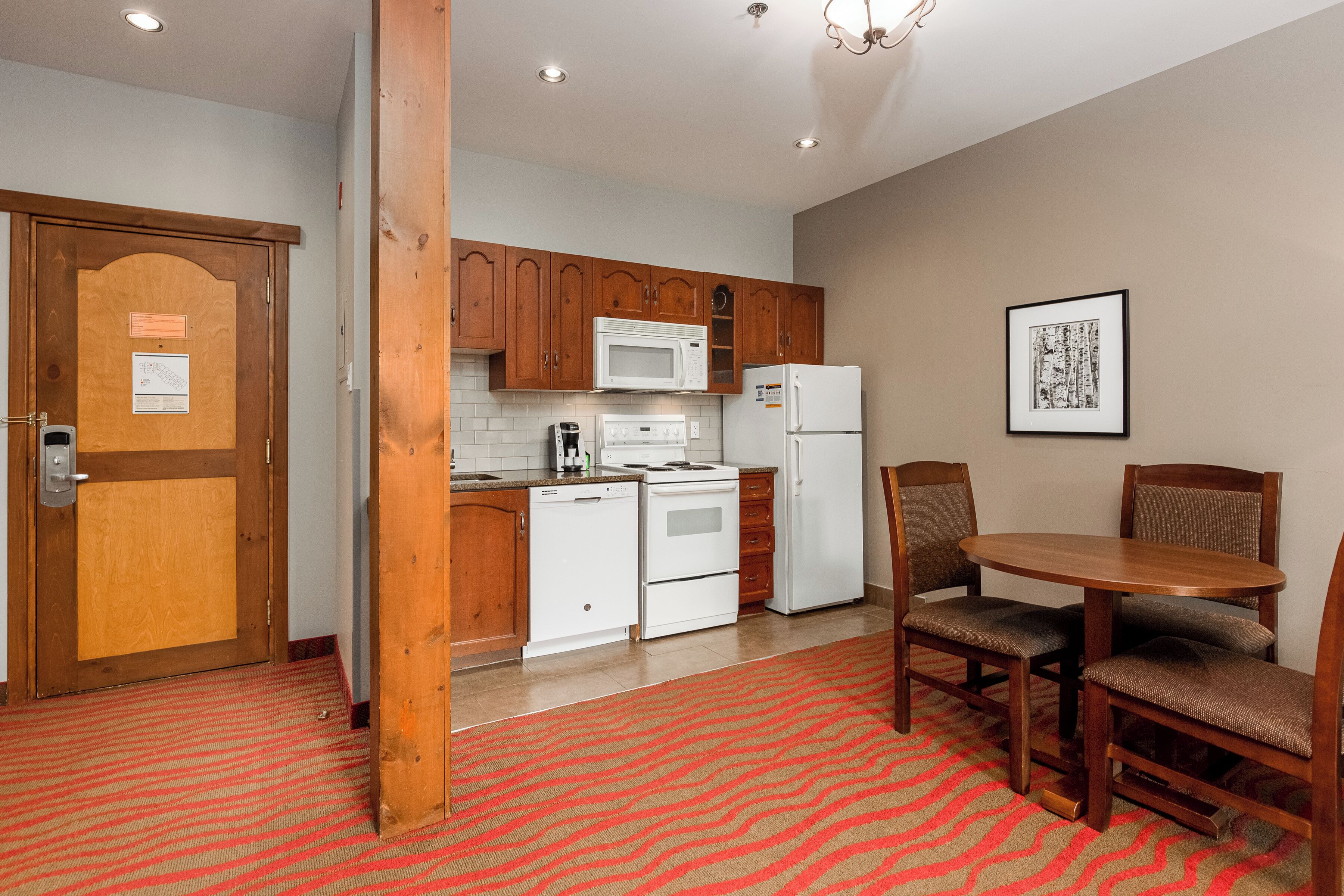 A compact kitchen with wooden cabinets, a microwave, a stove, and a refrigerator. There is a dining table with chairs and a red patterned carpet.