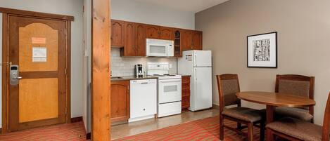 A compact kitchen with wooden cabinets, a microwave, a stove, and a refrigerator. There is a dining table with chairs and a red patterned carpet.