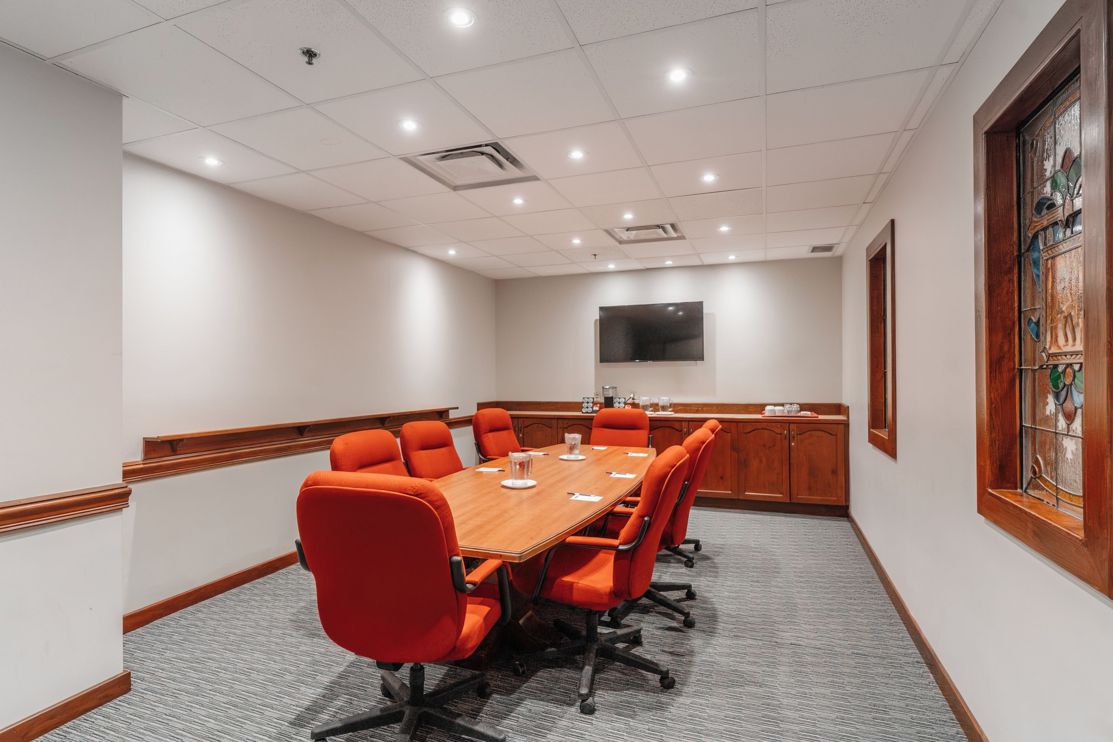 A conference room with a long table, multiple chairs, a flat-screen TV, and a stained glass window.
