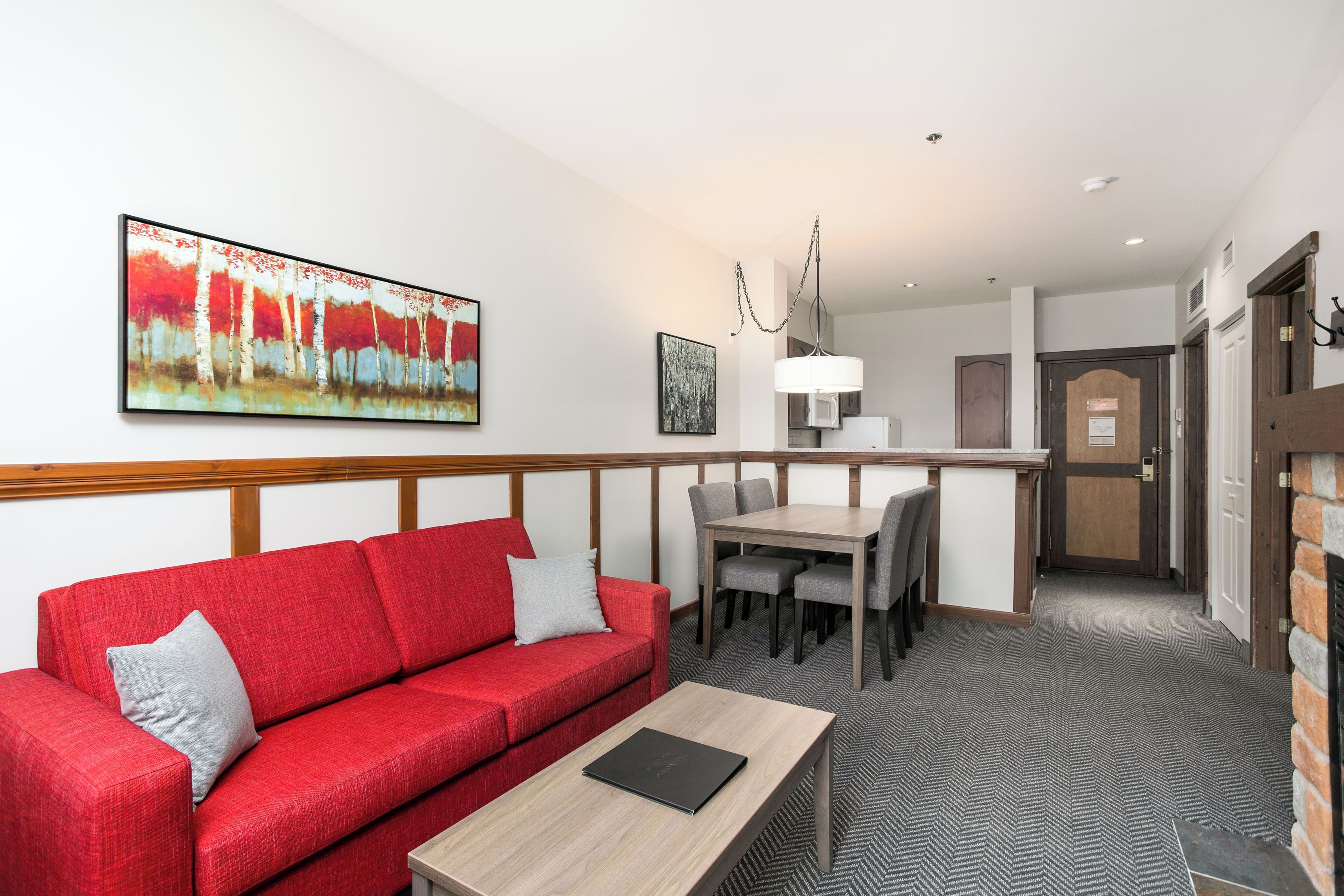 A hotel room with a red sofa, a wooden coffee table, a dining table with chairs, a painting on the wall, and a door leading to another room.