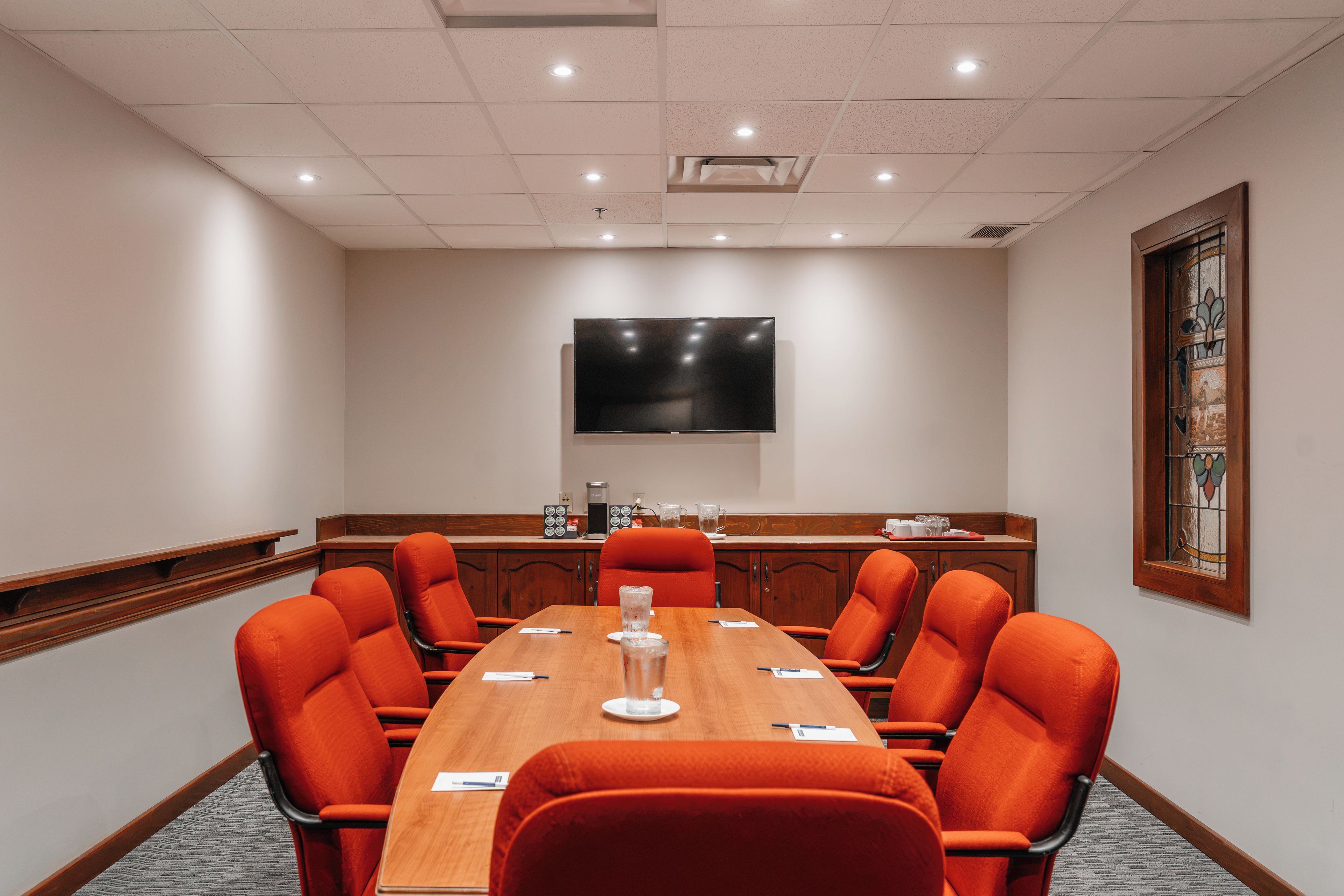 A conference room with a long table, orange chairs, a flat-screen TV on the wall, and a stained glass window.