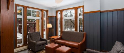 Una sala de estar acogedora con un sofá de cuero marrón, dos sillones y un reposapiés a juego. Amplias ventanas dejan entrar luz natural y ofrecen vistas al exterior.