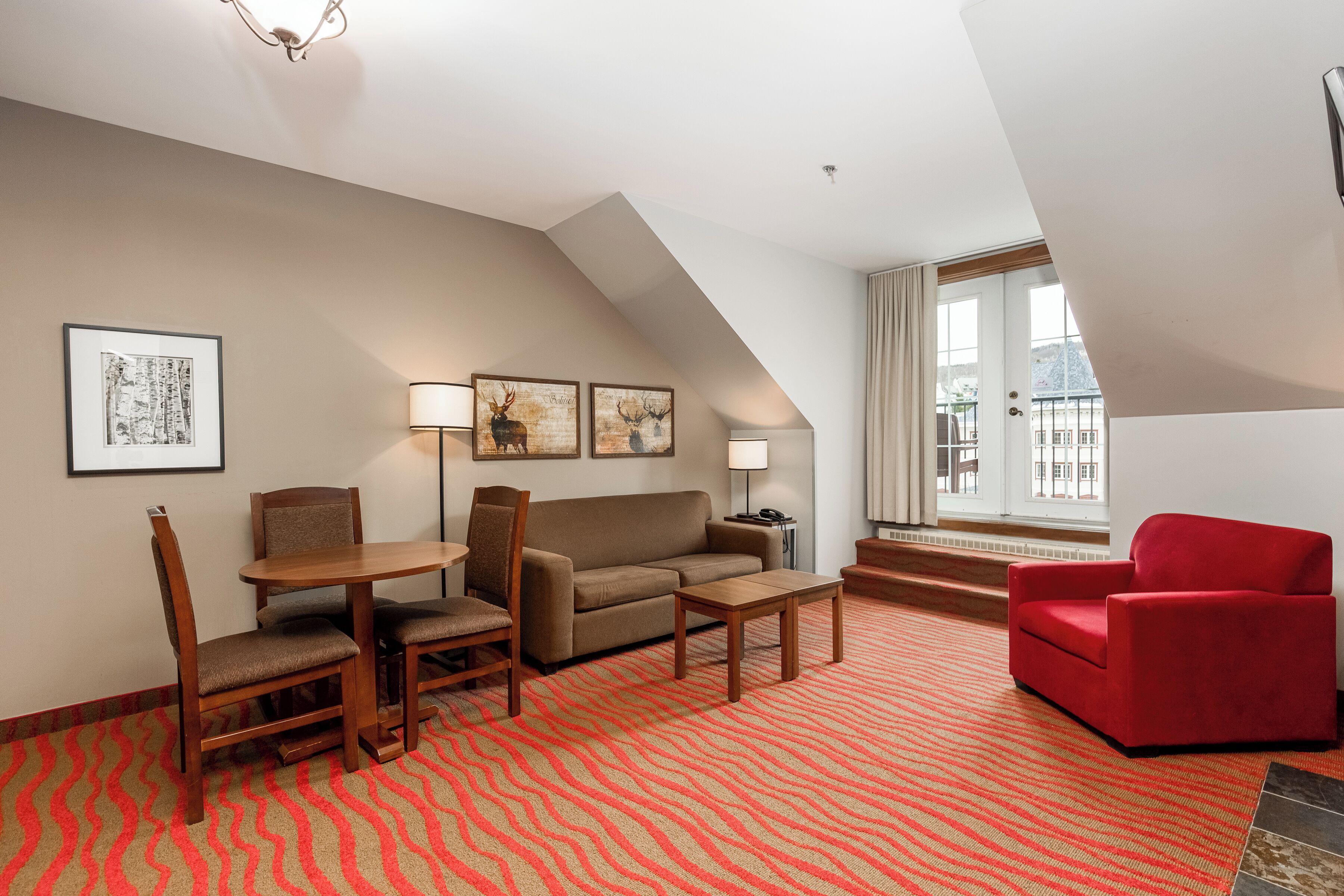 A modern living room with a sofa, two chairs, a coffee table, and a red armchair. There are framed pictures on the wall, a lamp, and a window with a view of buildings.