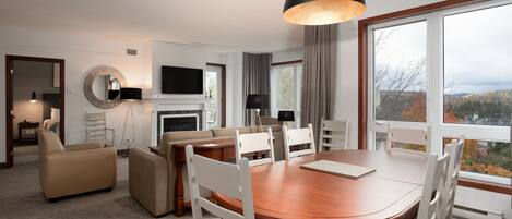 A modern hotel room with a dining area, a sofa set, a fireplace, and a large window with a city view.