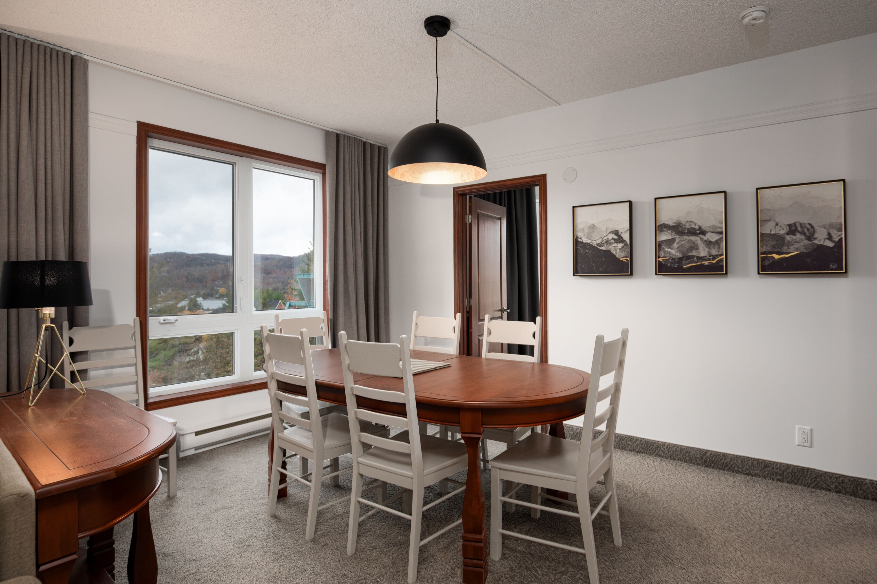 A dining area with a wooden table and white chairs, a pendant light, and a view of the outdoors through large windows.