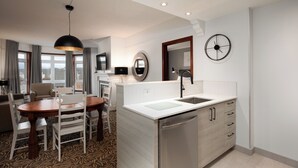 A modern kitchen with a dishwasher, a dining area with a wooden table and chairs, a large window with curtains, and a clock on the wall. - Sommet des Neiges (Mont-Tremblant)